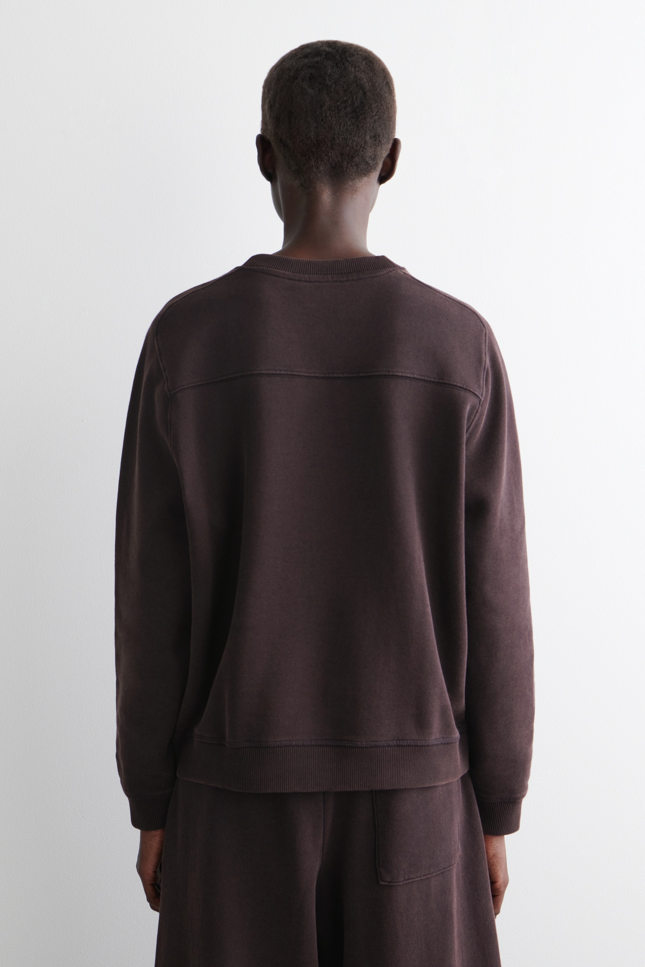 Agrandir l'image: Un sweat-shirt marron foncé est présenté de dos, avec un col rond, des poignets côtelés et un ourlet côtelé. Il présente un détail de couture horizontale sur le haut du dos et est assorti à un pantalon de survêtement marron foncé.