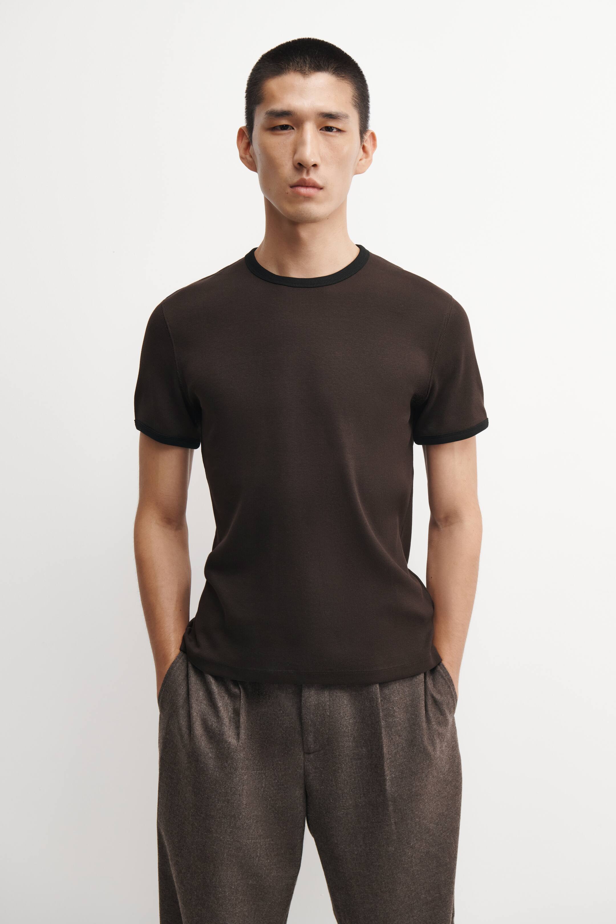 Ver imagen más grande: Se muestra una camiseta de algodón acanalado de corte ajustado en marrón oscuro, vista de frente, con cuello redondo negro en contraste y ribetes negros a juego en los puños de las mangas cortas. La camiseta, de ajuste ceñido, cae recta sobre la cintura de un pantalón marrón texturizado, realzando su silueta estilizada.