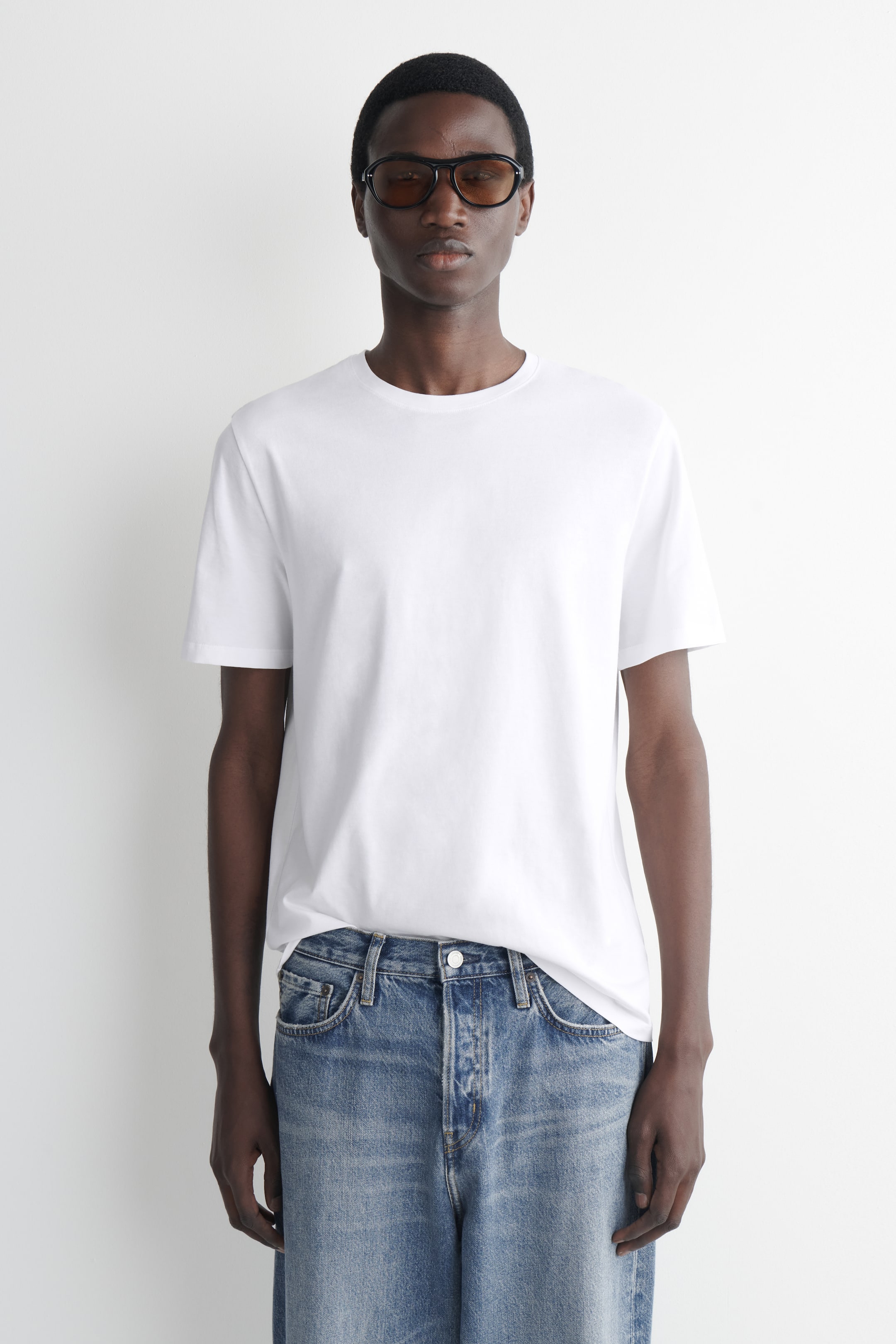 Größeres Bild anzeigen: Ein weißes T-Shirt wird getragen, nach vorne gerichtet, mit Rundhalsausschnitt und kurzen Ärmeln. Der vordere Saum ist teilweise in blaue Jeans gesteckt.