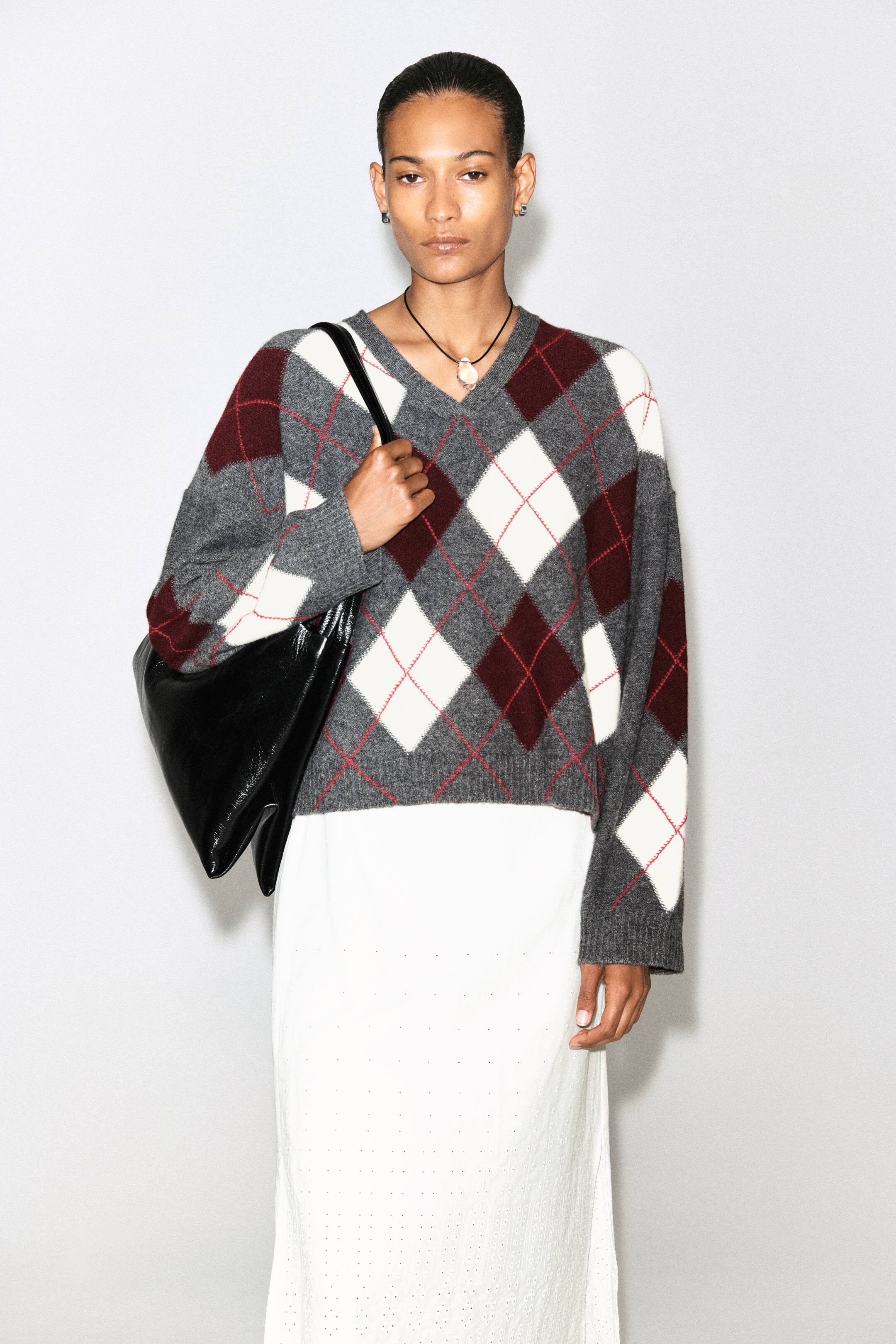 Größeres Bild ansehen: Ein grauer, bordeauxfarbener und weißer Argyle-Pullover mit roten Umrandungsfäden wird vom Model getragen. Dieser verkürzte Pullover verfügt über einen V-Ausschnitt und Rippbündchen und ist über einem weißen Rock kombiniert. Das Model trägt außerdem eine schwarze Schultertasche über dem Pullover.