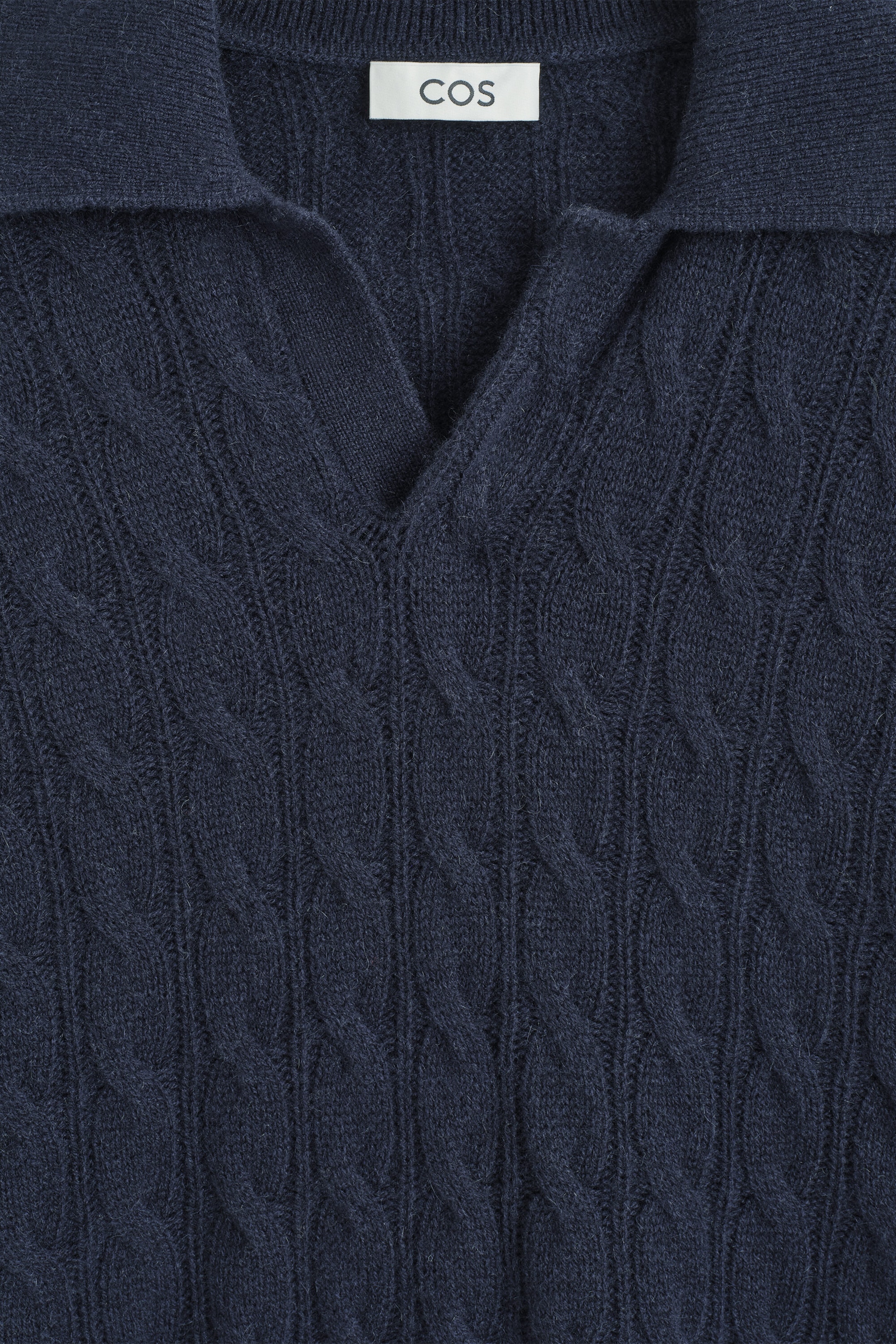A cable-knit cashmere polo sweater in dark navy blue is shown in a close-up front view, highlighting its soft, textured fabric and intricate cable-knit pattern. The garment features an open, buttonless polo collar with a V-neckline and a white woven label displaying the brand name COS inside the collar.