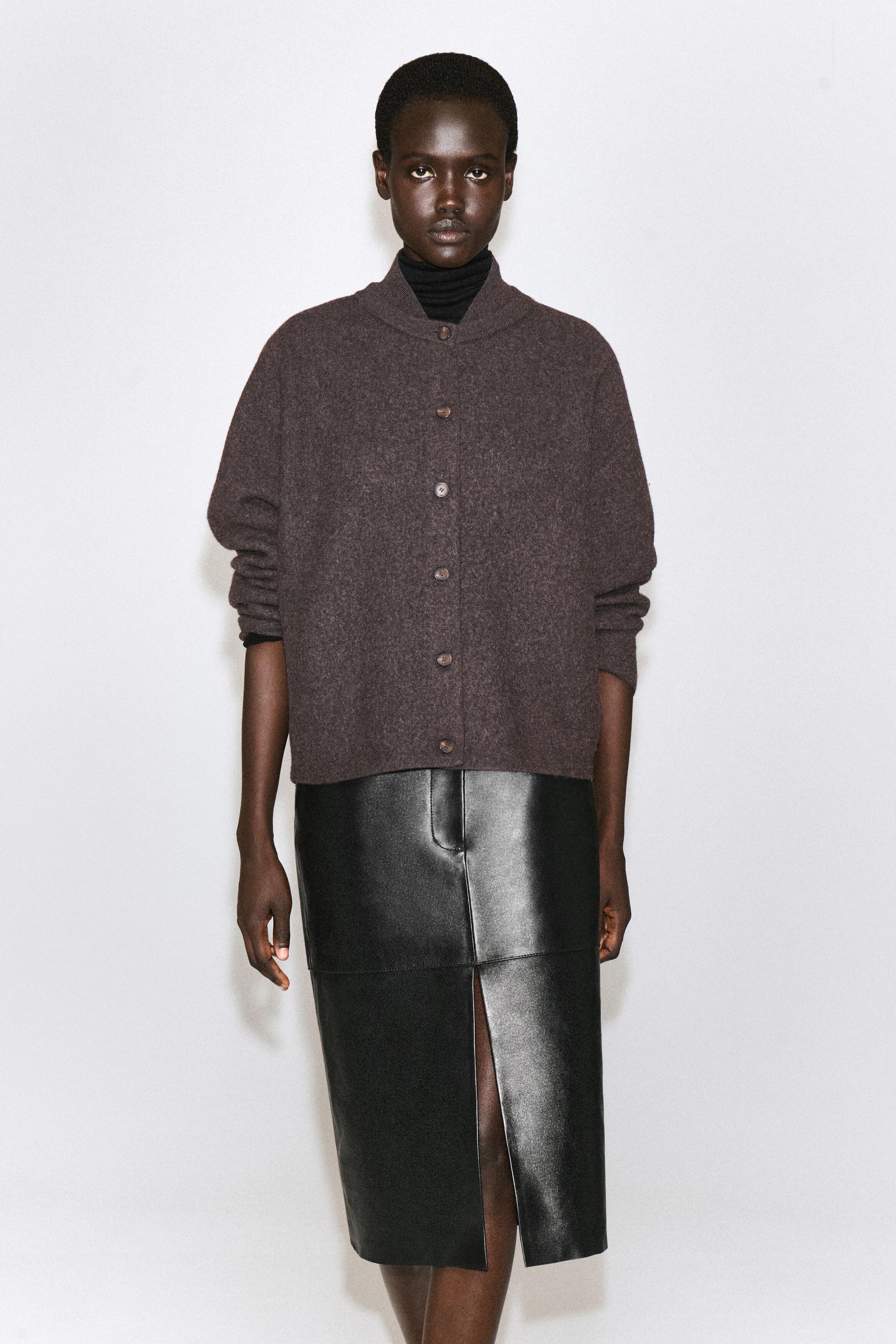 Vue de face d’un cardigan en laine bouillie brun foncé présentant une silhouette doucement arrondie, une fermeture à boutons sur le devant et un col de style bomber à effet superposé. Le cardigan est porté par-dessus un col roulé noir et agencé avec une jupe midi en cuir noir. Vue de face d’un cardigan en laine bouillie brun foncé présentant une silhouette doucement arrondie, une fermeture à boutons sur le devant et un col de style bomber à effet superposé. Le cardigan est porté par-dessus un col roulé noir et agencé avec une jupe midi en cuir noir.