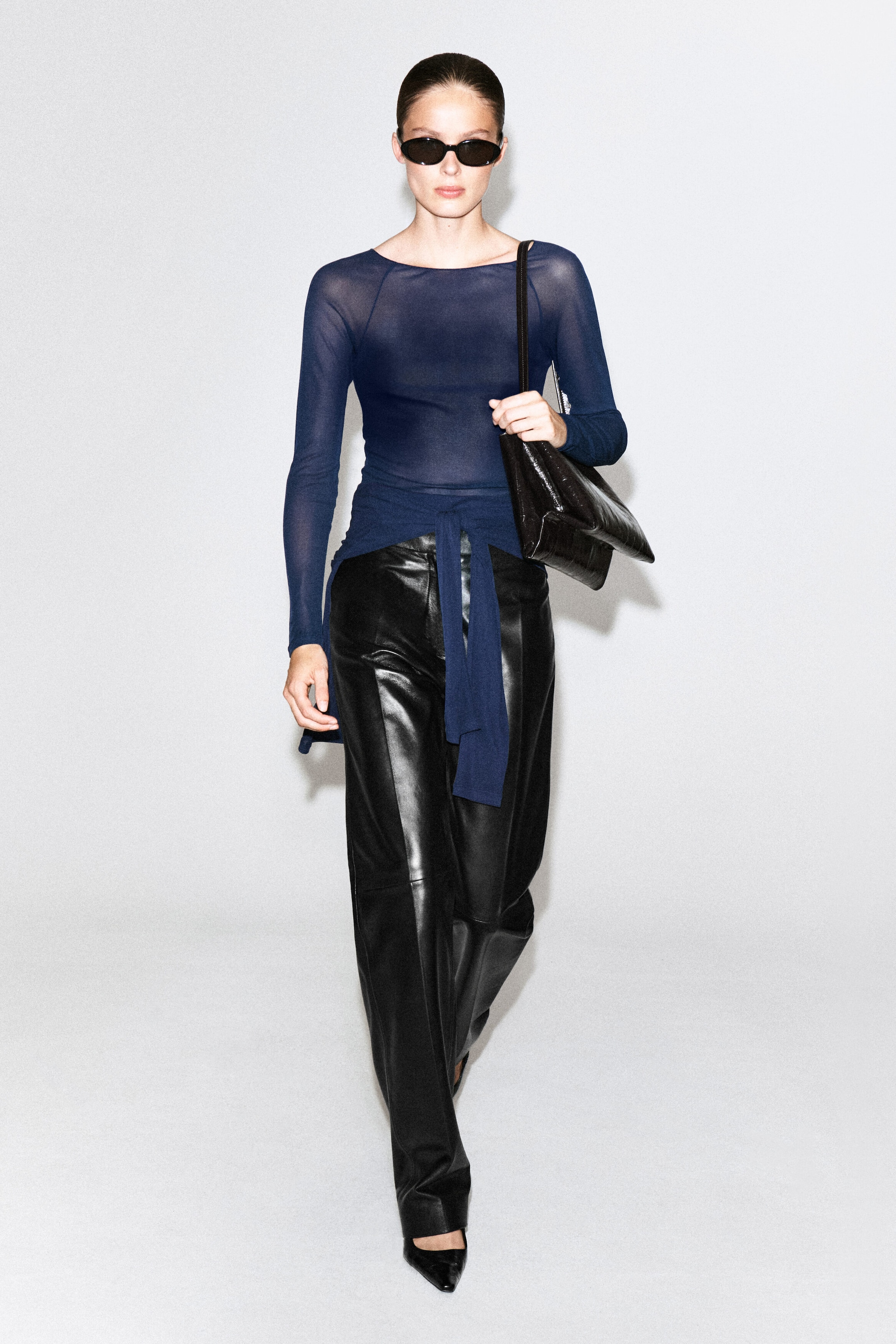 Le haut en soie côtelée ajusté, bleu marine, est porté par le mannequin. Il présente une texture légèrement transparente, des manches longues et une ceinture assortie en tissu drapée à la taille. Le haut est agencé avec un pantalon noir en cuir à jambe large et un sac à bandoulière noir.