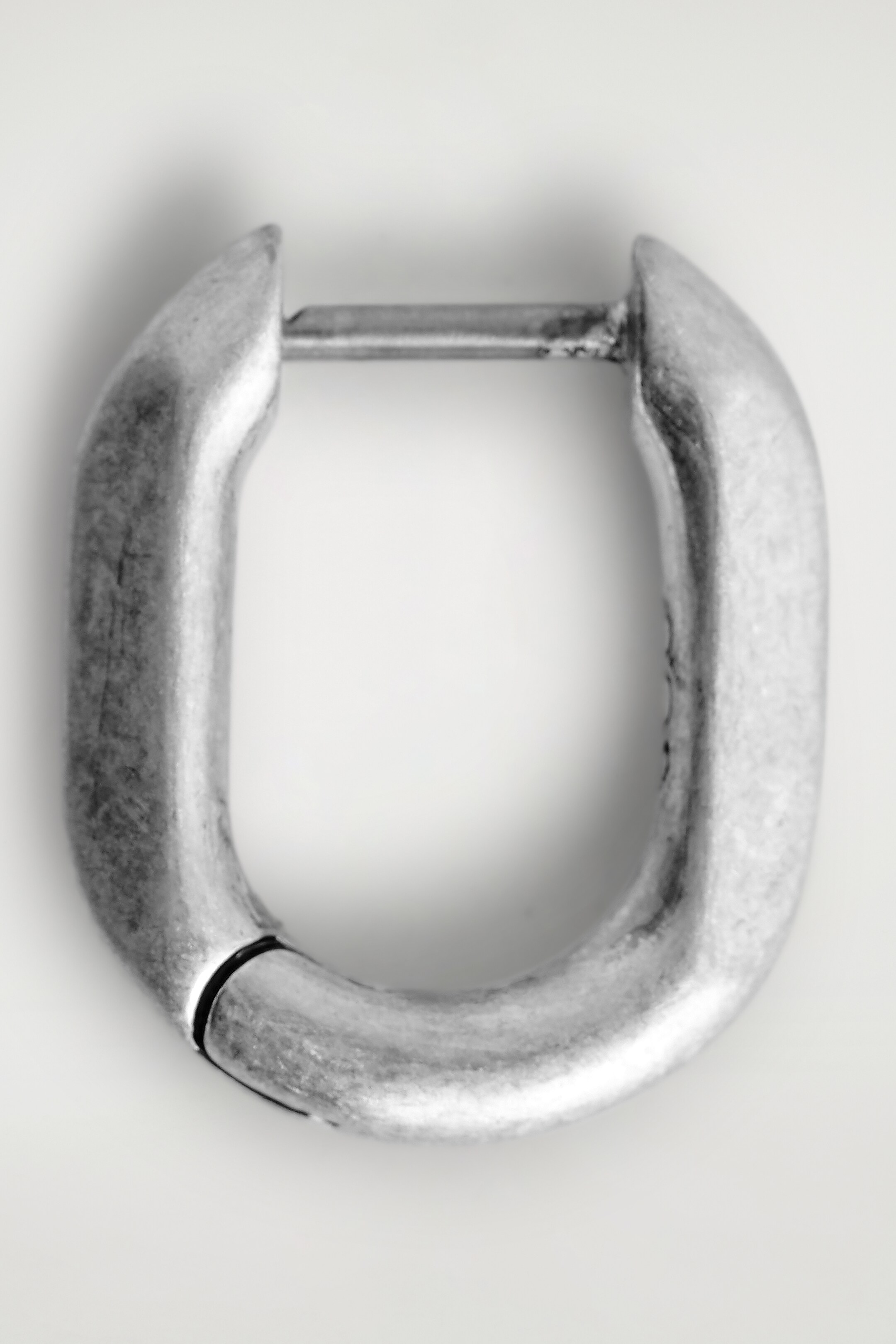Agrandir l'image: Une seule boucle d'oreille en forme de cerceau argenté, orientée vers l'avant, met en valeur sa forme rectangulaire allongée et arrondie. Elle présente une finition mate et vieillie avec une fermeture à charnière visible à sa courbe inférieure et une fermeture à épingle à sa courbe supérieure.