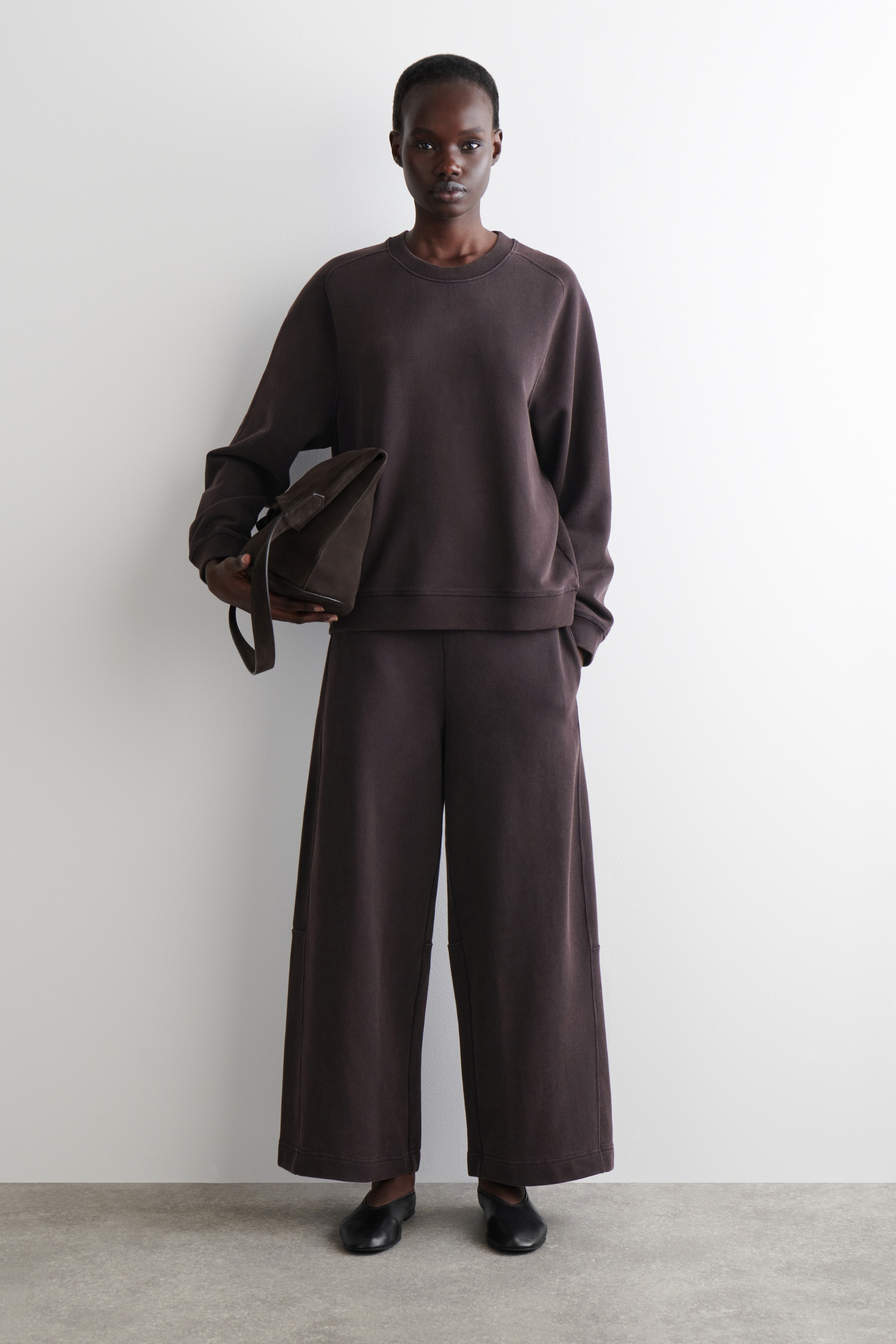 Agrandir l'image: Un ensemble assorti marron foncé à coupe décontractée est présenté, comprenant un sweat-shirt à col rond avec des épaules tombantes, des poignets côtelés et un ourlet côtelé, associé à un pantalon large et court avec des détails de coutures verticales. Un sac fourre-tout marron foncé est tenu, complétant le look avec des chaussures plates noires.