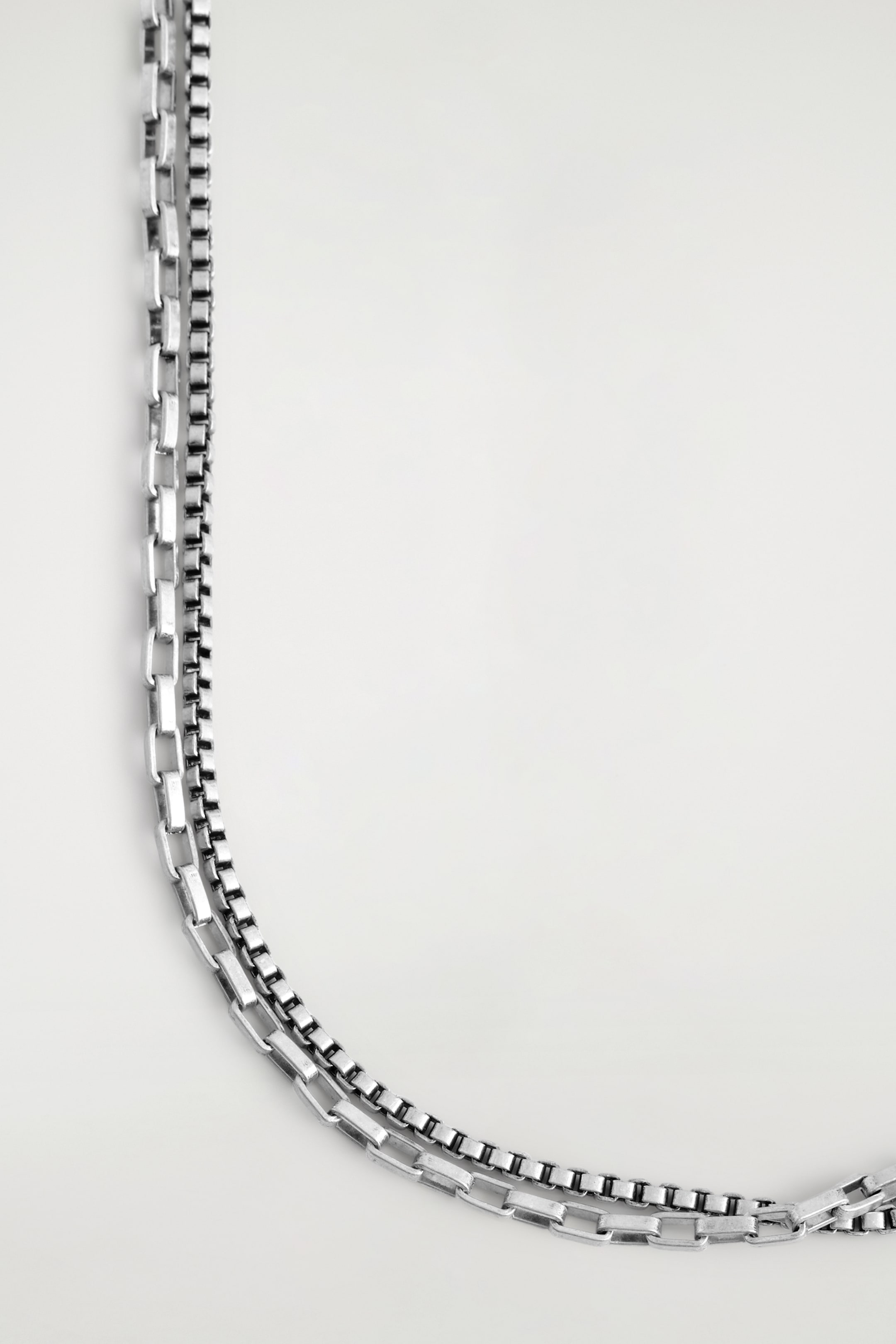 Visualizza immagine più grande: Una collana color argento mostra il suo design a doppio strato, caratterizzato da una classica catena a maglie quadrate e una catena a maglie rettangolari allungate.