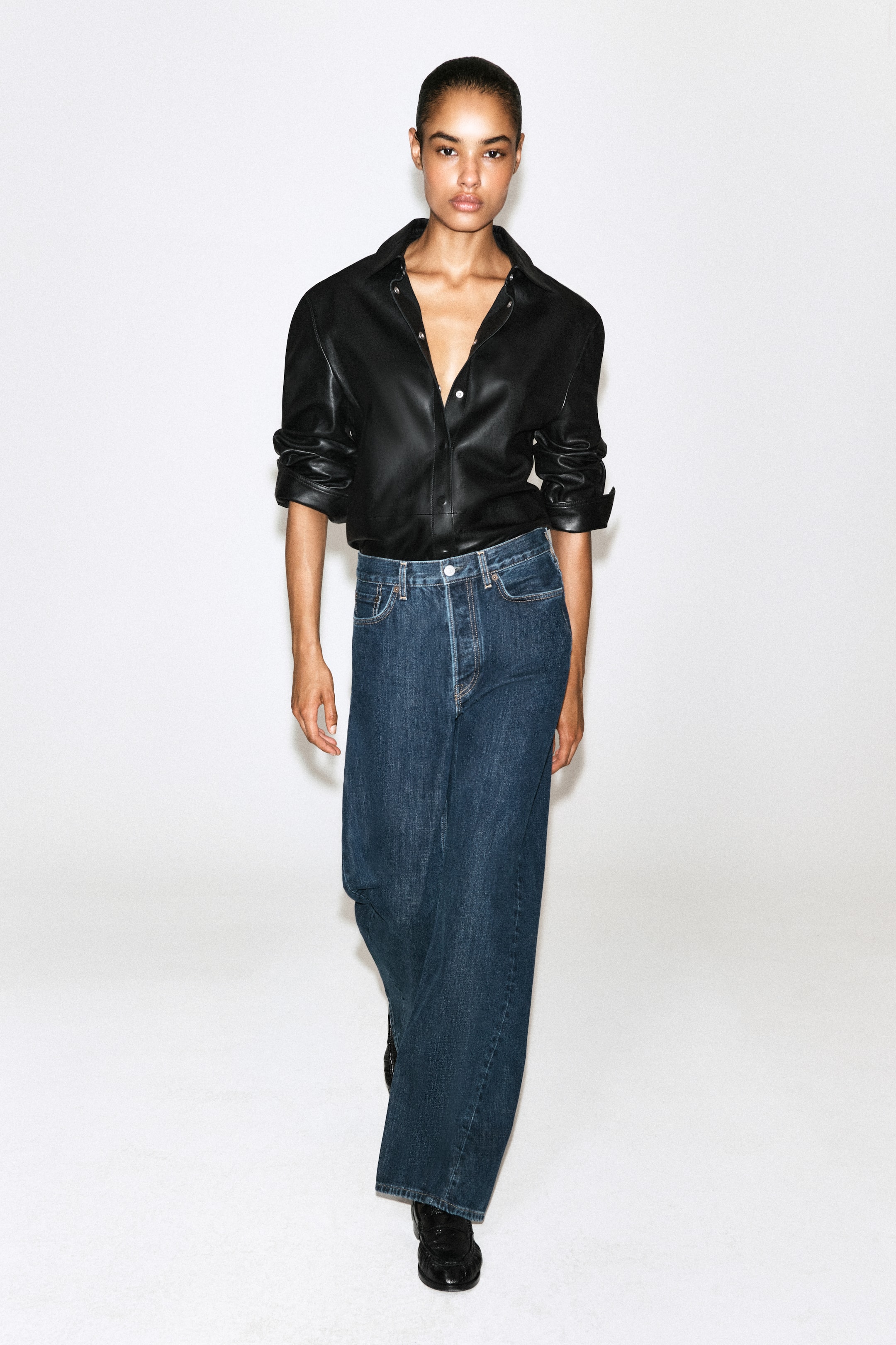 Se større billede: Forfra bærer modellen højtaljede jeans med brede ben i en mørkeblå vask og en afslappet pasform. Jeansene styles med en nedstukket sort læderskjorte med oprullede ærmer og en åben krave.