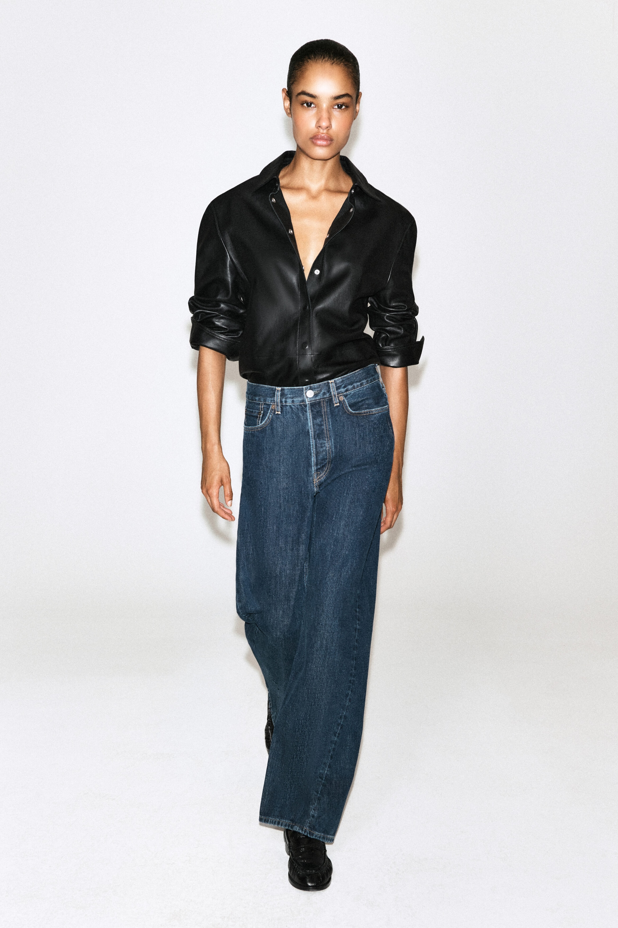 Facing forward, the model is wearing high-waisted, wide-leg jeans in a dark blue wash with a relaxed fit. The jeans are paired with a tucked-in black leather shirt featuring rolled-up sleeves and an unbuttoned collar. Facing forward, the model is wearing high-waisted, wide-leg jeans in a dark blue wash with a relaxed fit. The jeans are paired with a tucked-in black leather shirt featuring rolled-up sleeves and an unbuttoned collar.
