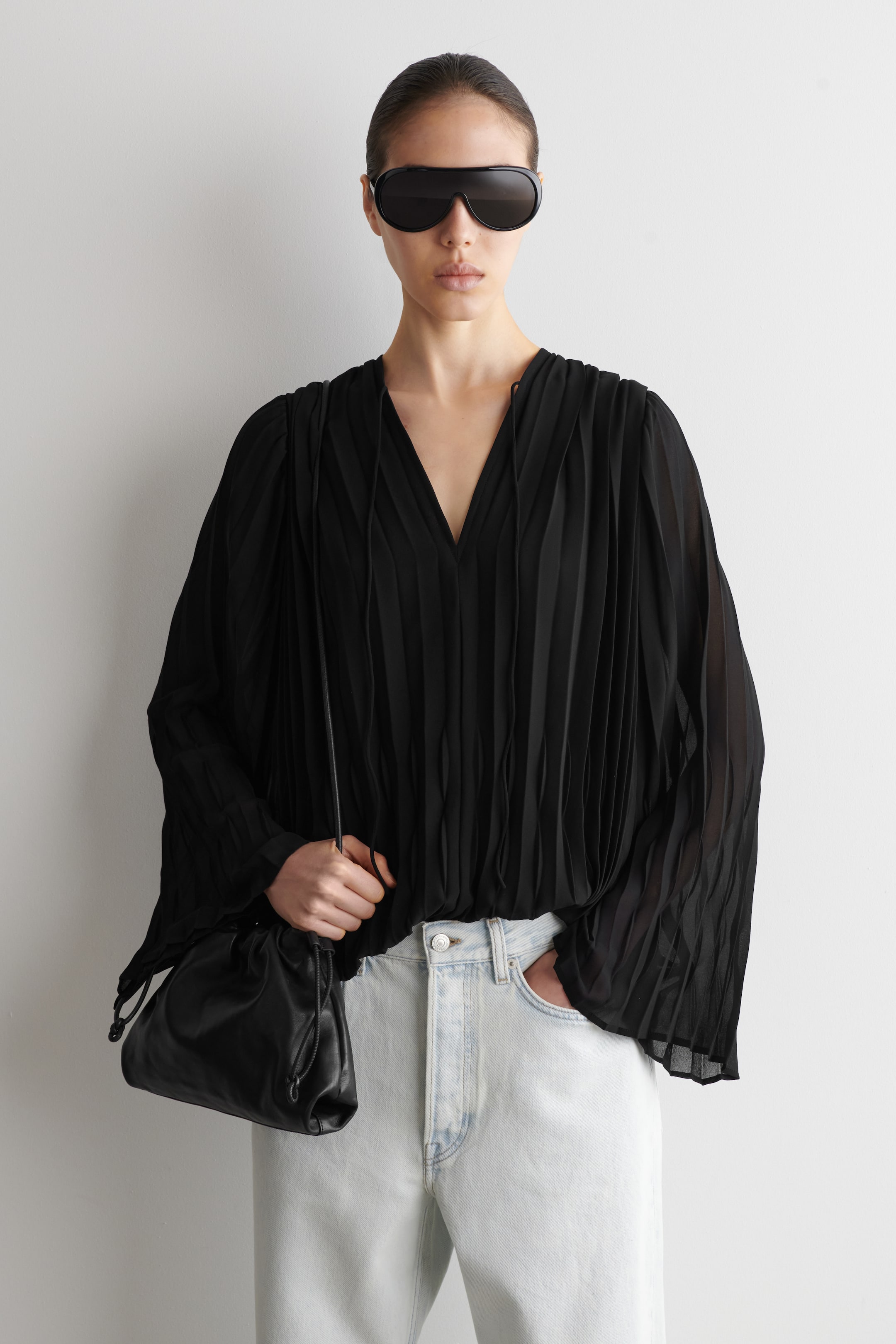 Größeres Bild ansehen: Eine schwarze plissierte Bluse mit V-Ausschnitt und Bindebändern sowie langen, lockeren Ärmeln. Der plissierte, halbtransparente Stoff bildet eine übergroße Silhouette, wobei eine Seite des Saums in eine hell gewaschene Jeans gesteckt ist.