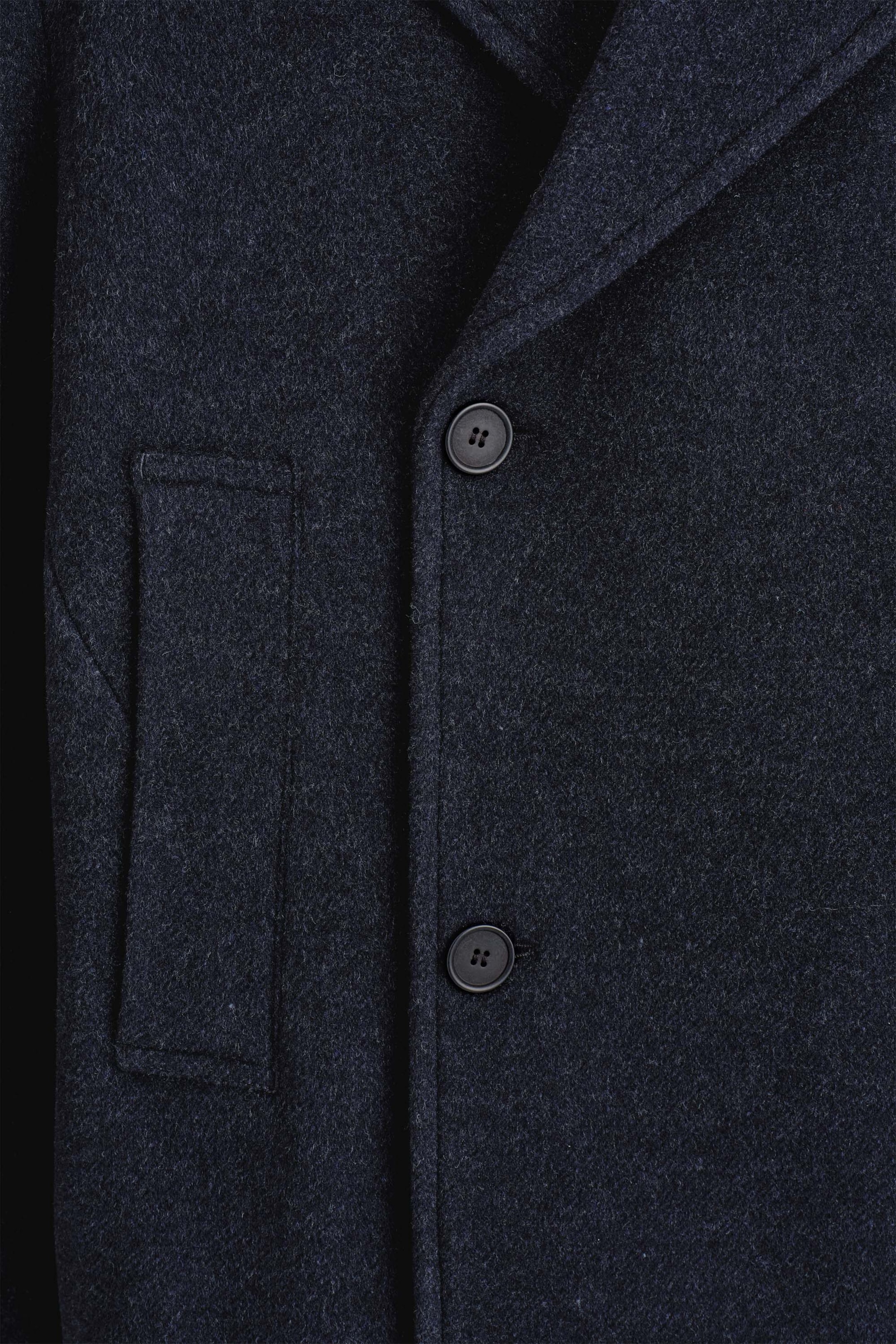 A close-up of the double-breasted wool short pea coat highlights the dark navy, textured wool fabric, showing two black buttons and a vertical welt pocket on the front. A section of the structured notch lapel extends from the collar area.