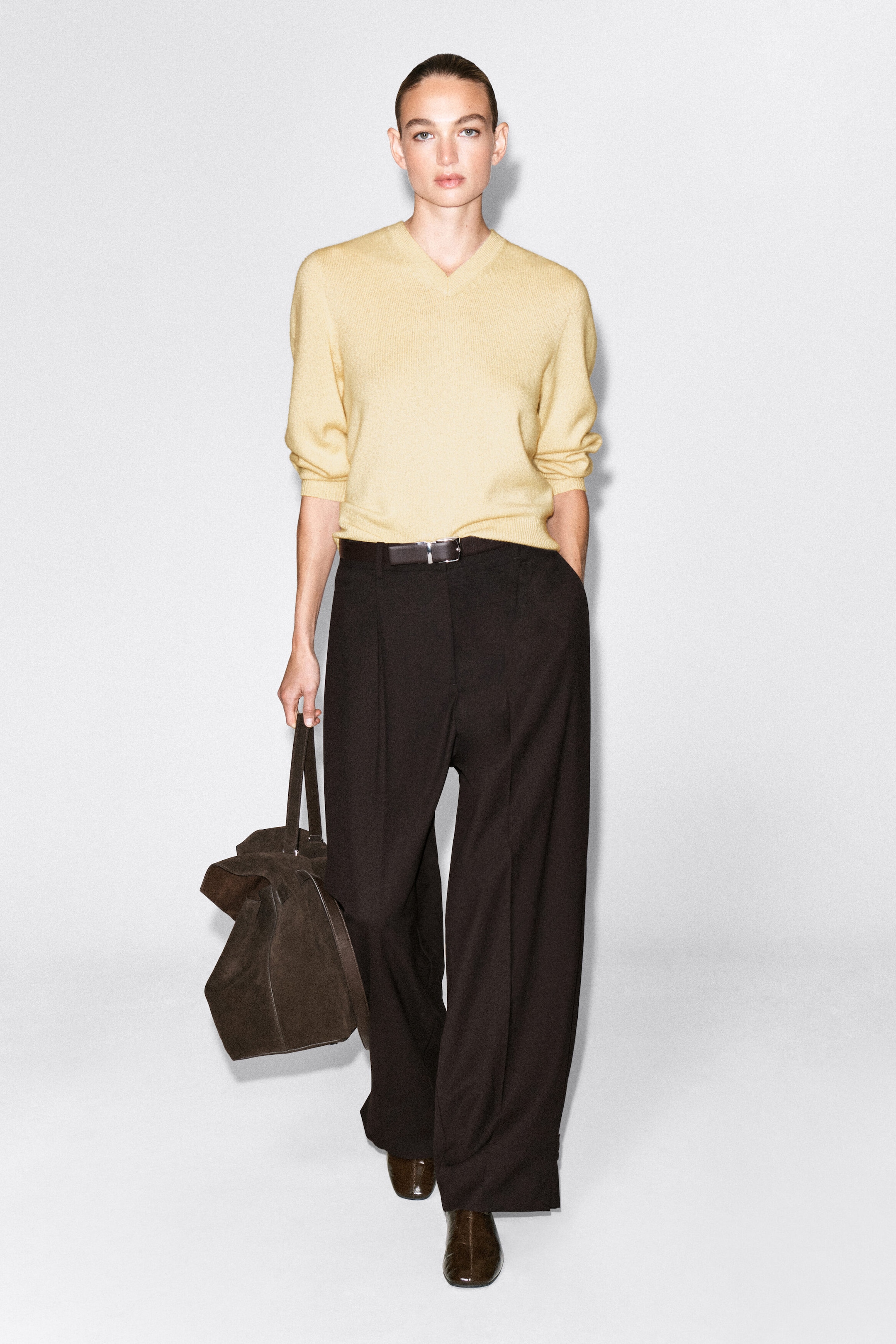 Das Model trägt einen hellgelben Strickpullover mit V-Ausschnitt, der in eine dunkelbraune Wide-Leg-Hose gesteckt ist, die mit einem schmalen Gürtel fixiert wird. Eine dunkelbraune Wildlederhandtasche wird in der rechten Hand des Models getragen und rundet das Outfit ab. Das Model trägt einen hellgelben Strickpullover mit V-Ausschnitt, der in eine dunkelbraune Wide-Leg-Hose gesteckt ist, die mit einem schmalen Gürtel fixiert wird. Eine dunkelbraune Wildlederhandtasche wird in der rechten Hand des Models getragen und rundet das Outfit ab.