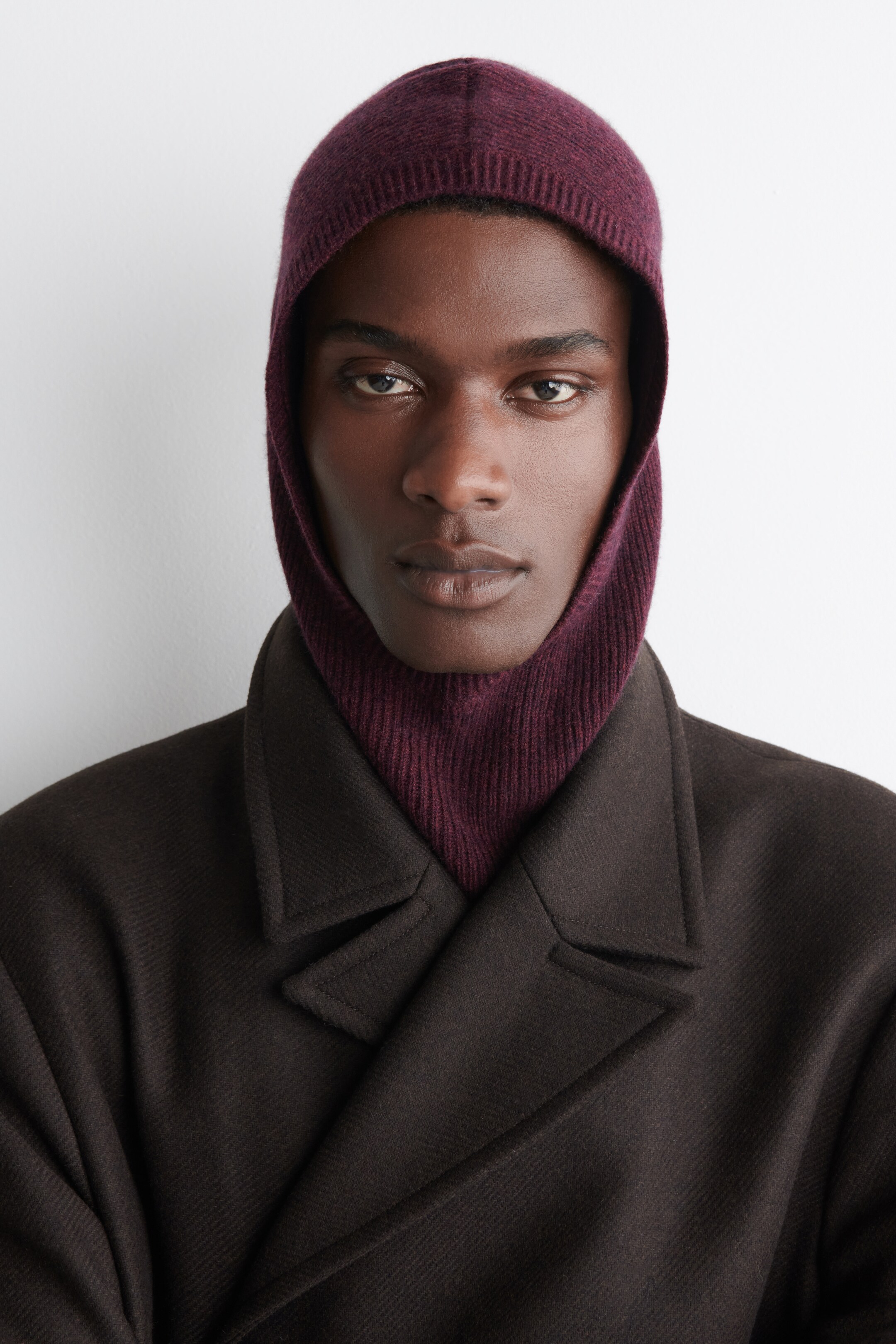 View larger image: A deep reddish-purple knitted balaclava is covering the head and neck, featuring a ribbed section down the front, and is being styled with a dark brown coat with wide lapels.