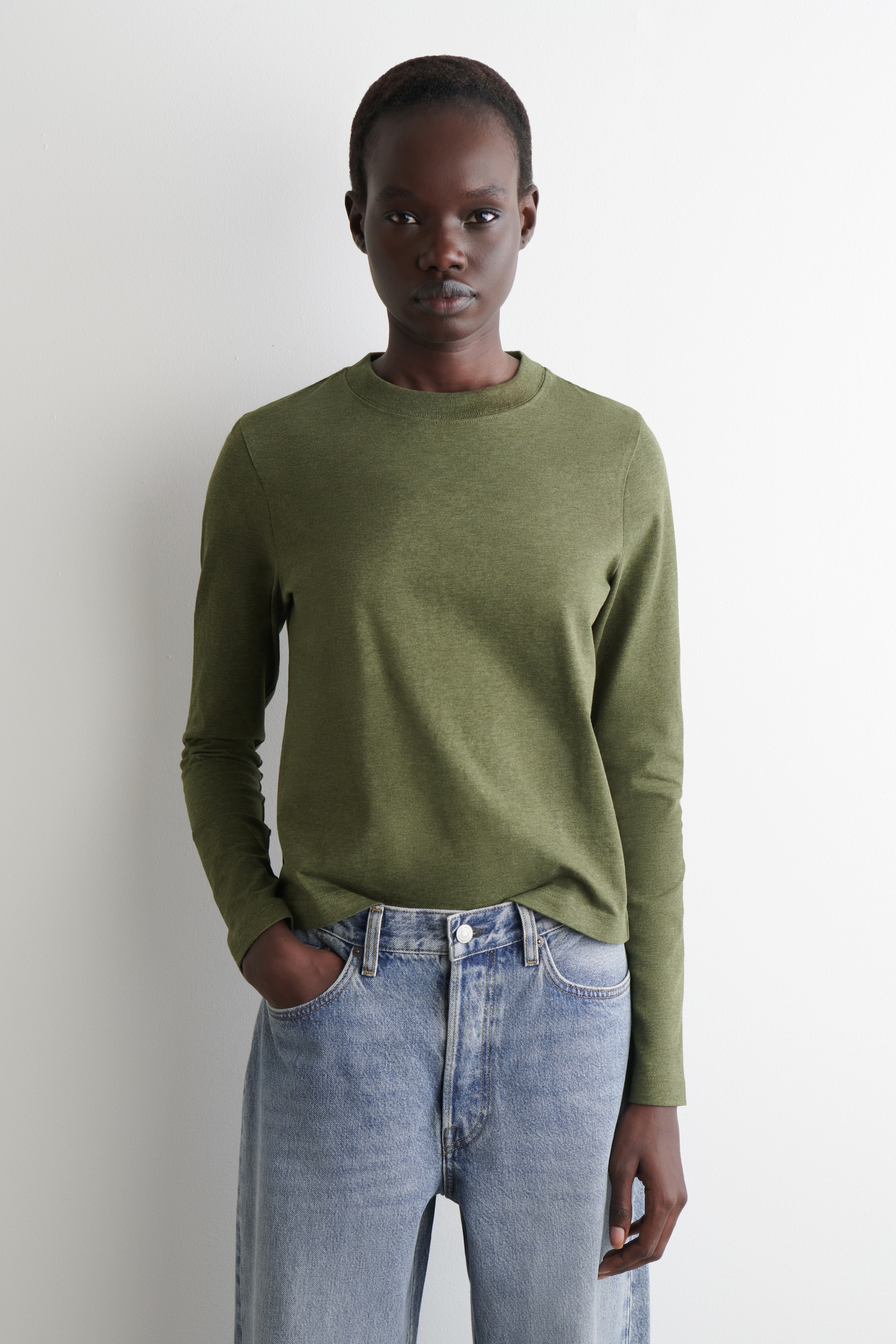 Vezi imaginea mai mare: Un tricou cu mâneci lungi de culoare verde măsliniu este purtat, orientat spre FAȚĂ. Acesta are un guler rotund, mâneci lungi și o textură discretă melanj, prezentând o potrivire îngrijită și este parțial băgat în blugi de culoare albastru deschis.