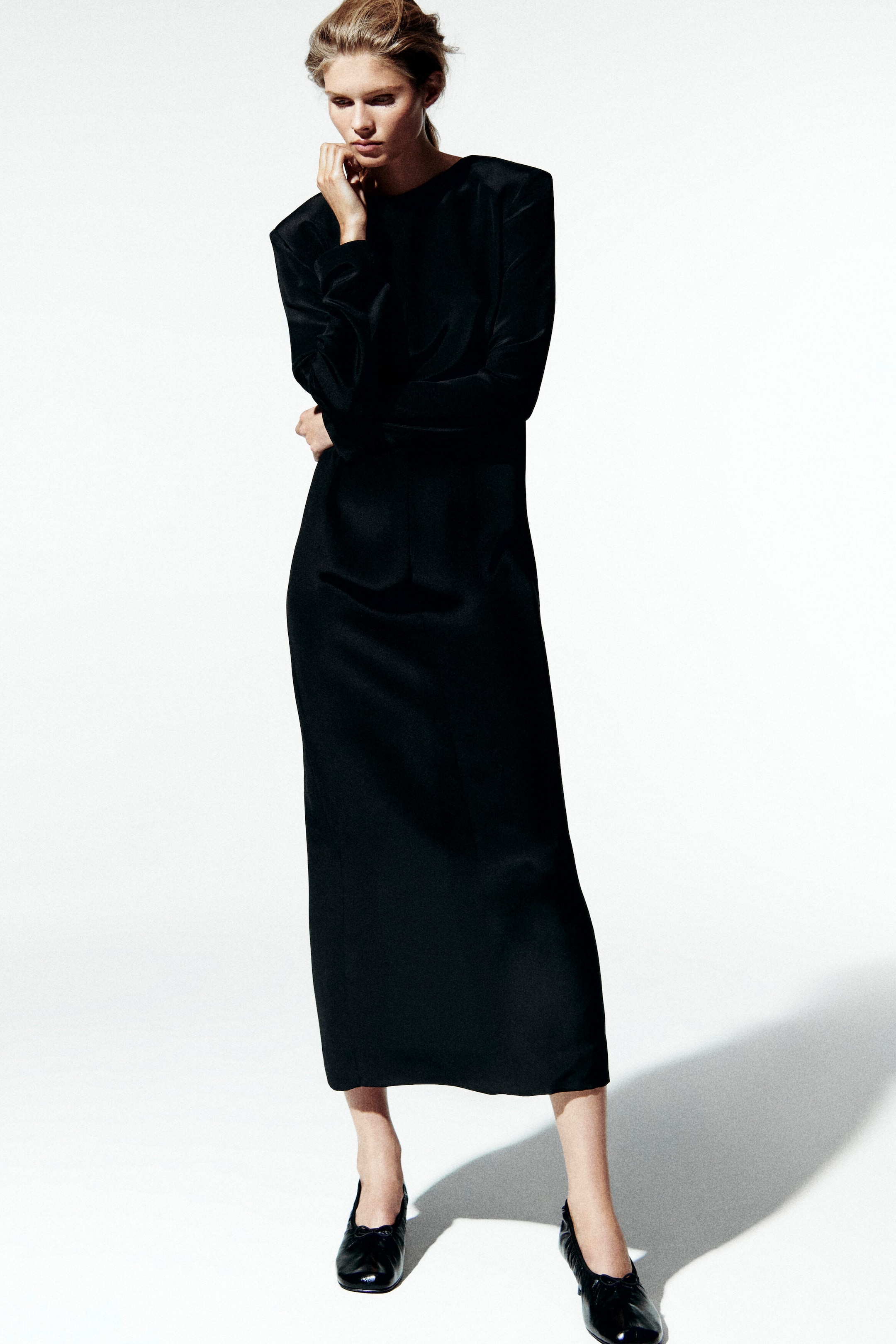 The button-detailed silk crepe maxi dress is shown from the front, featuring a high round neckline, structured shoulders, and long sleeves. This black dress drapes in a straight column silhouette to an ankle-grazing length and is paired with black flat shoes. The button-detailed silk crepe maxi dress is shown from the front, featuring a high round neckline, structured shoulders, and long sleeves. This black dress drapes in a straight column silhouette to an ankle-grazing length and is paired with black flat shoes.