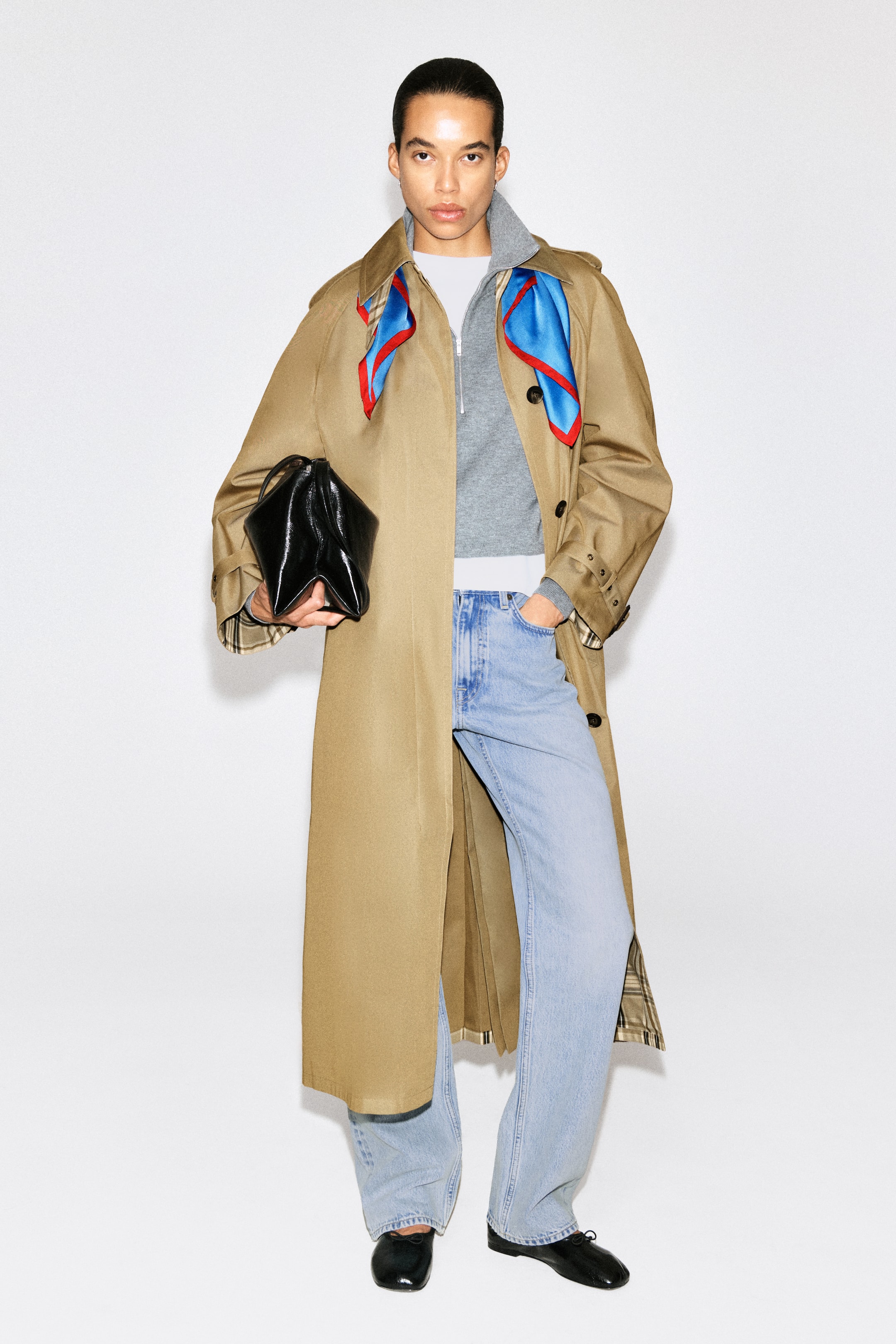 Преглед на по-голямо изображение: CHECK-DETAIL BELTED COTTON TRENCH COAT е представено отпред, с лека кафява памучна материя и класическа отложна яка. Палто̀то е стилизирано отворено върху сив суитшърт с качулка и шал с синьо-червен десен, а вътрешната му част показва каре подплата в края и по маншетите на ръкавите. Регулируеми каишки на маншетите подчертават дългите ръкави.