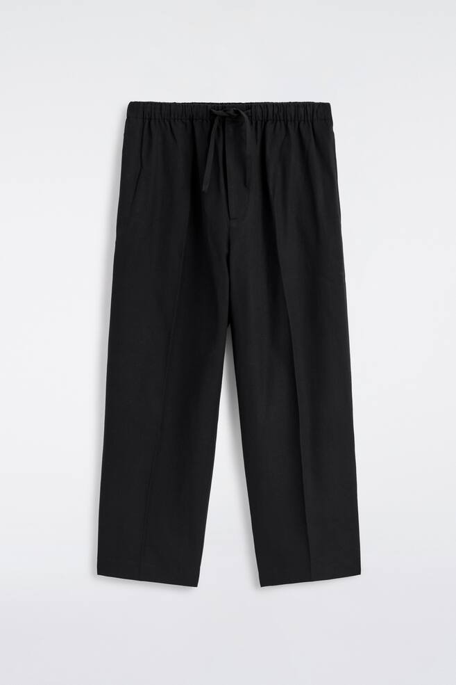 Schwarze Hosen werden von vorne gezeigt und verfügen über einen elastischen Bund mit einem Kordelzug in der Mitte. Sie sind in einer Barrel-Leg-Form gestaltet, mit dezenten Seitentaschen und feinen vertikalen Falten, die an der Vorderseite jedes Beins herunterlaufen. - 1