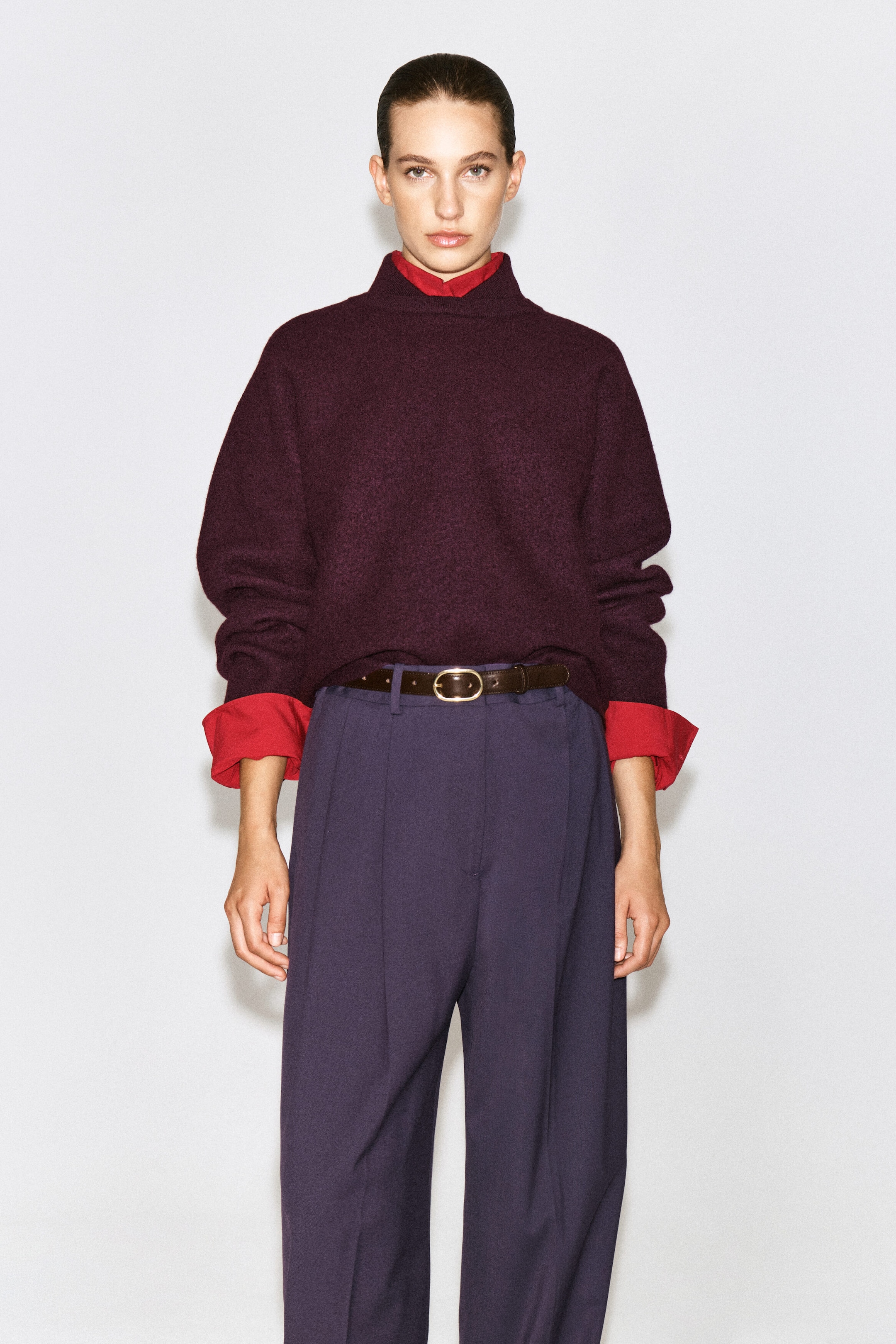 Un chandail en laine bouillie de couleur prune foncée est porté par le mannequin, superposé à une chemise rouge éclatante dont le col et les poignets retroussés sont visibles. Le chandail à col montant, à coupe décontractée, est rentré dans un pantalon large violet foncé maintenu par une ceinture en cuir brun. Un chandail en laine bouillie de couleur prune foncée est porté par le mannequin, superposé à une chemise rouge éclatante dont le col et les poignets retroussés sont visibles. Le chandail à col montant, à coupe décontractée, est rentré dans un pantalon large violet foncé maintenu par une ceinture en cuir brun.