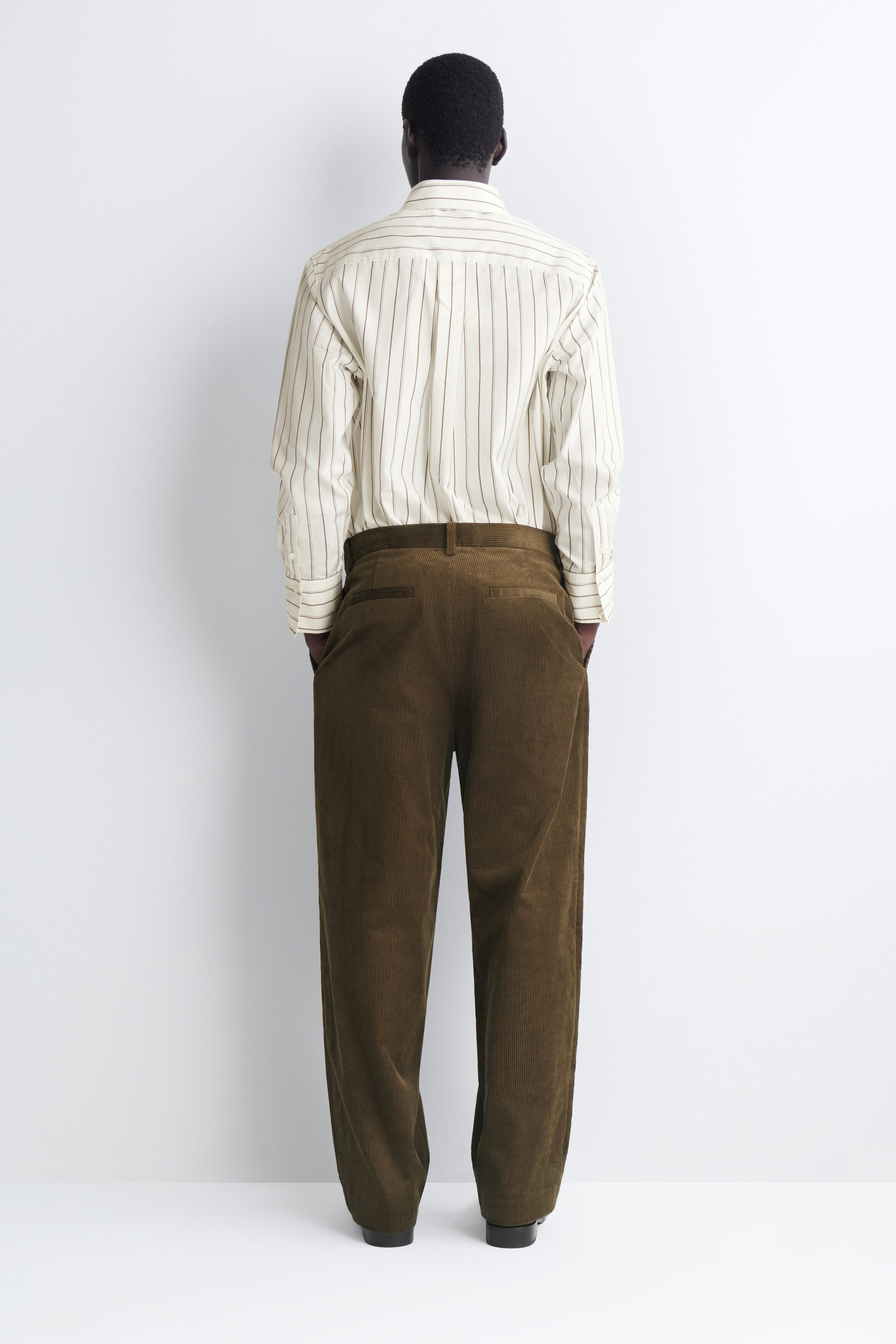 The Pleated Relaxed-Fit Tapered Corduroy Pants in olive brown are shown from the back, showcasing a relaxed, tapered fit. The pants feature belt loops and rear welt pockets and are styled with a tucked-in striped long-sleeve shirt. The Pleated Relaxed-Fit Tapered Corduroy Pants in olive brown are shown from the back, showcasing a relaxed, tapered fit. The pants feature belt loops and rear welt pockets and are styled with a tucked-in striped long-sleeve shirt.