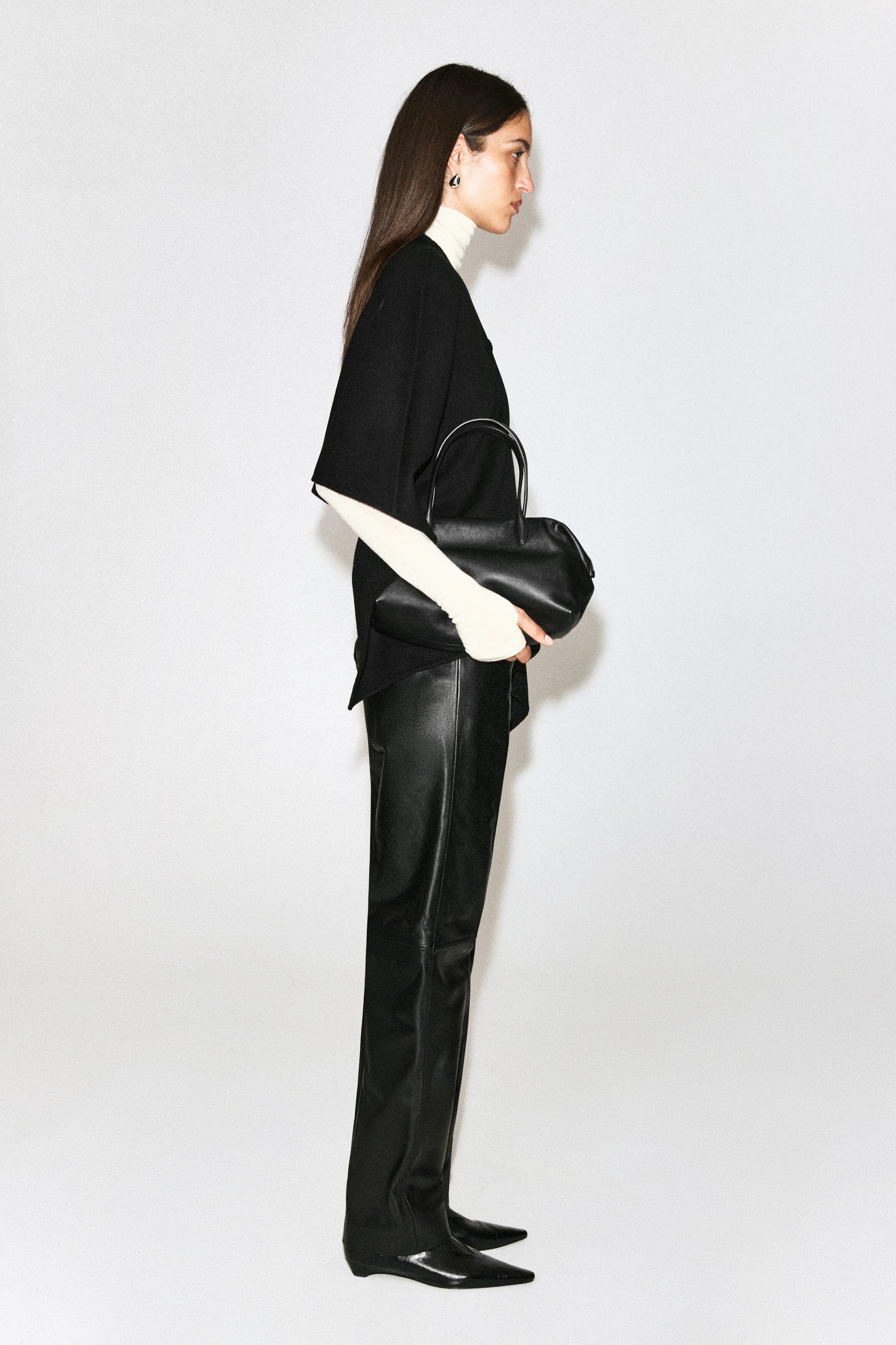 A black double-faced wool-blend cape is being worn by the model over a cream-coloured high-neck top. The cape features short, wide sleeves and is styled with black leather trousers, while a black leather handbag is being carried. A black double-faced wool-blend cape is being worn by the model over a cream-coloured high-neck top. The cape features short, wide sleeves and is styled with black leather trousers, while a black leather handbag is being carried.