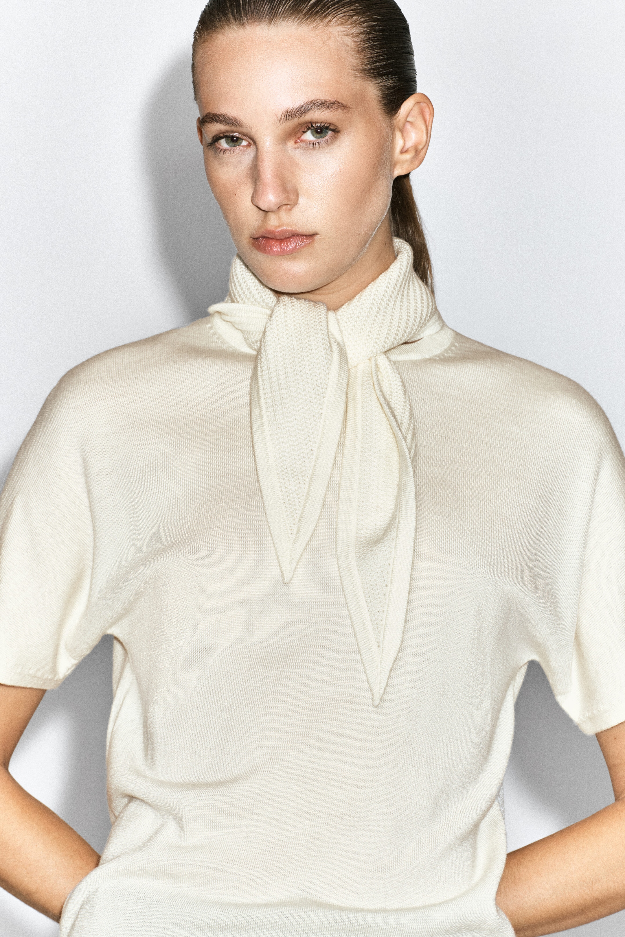 A cream-colored merino wool-blend T-shirt with short sleeves and an integrated matching knit scarf tied around the neck is worn by the model.