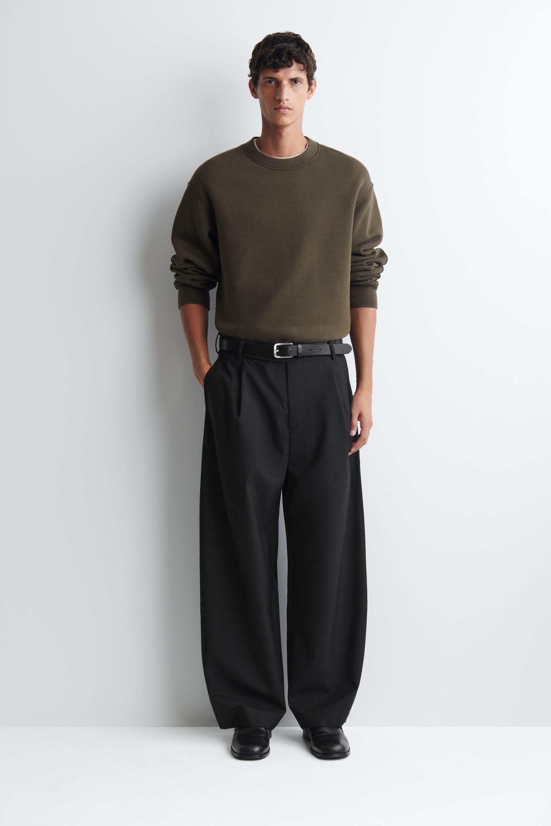 Le mannequin porte un sweatshirt en jersey de coton et laine vert olive, rentré dans un pantalon noir à jambes larges avec une ceinture. Le sweatshirt présente un col rond côtelé, des épaules tombantes et des poignets côtelés légèrement remontés sur les avant-bras. Le mannequin porte un sweatshirt en jersey de coton et laine vert olive, rentré dans un pantalon noir à jambes larges avec une ceinture. Le sweatshirt présente un col rond côtelé, des épaules tombantes et des poignets côtelés légèrement remontés sur les avant-bras.