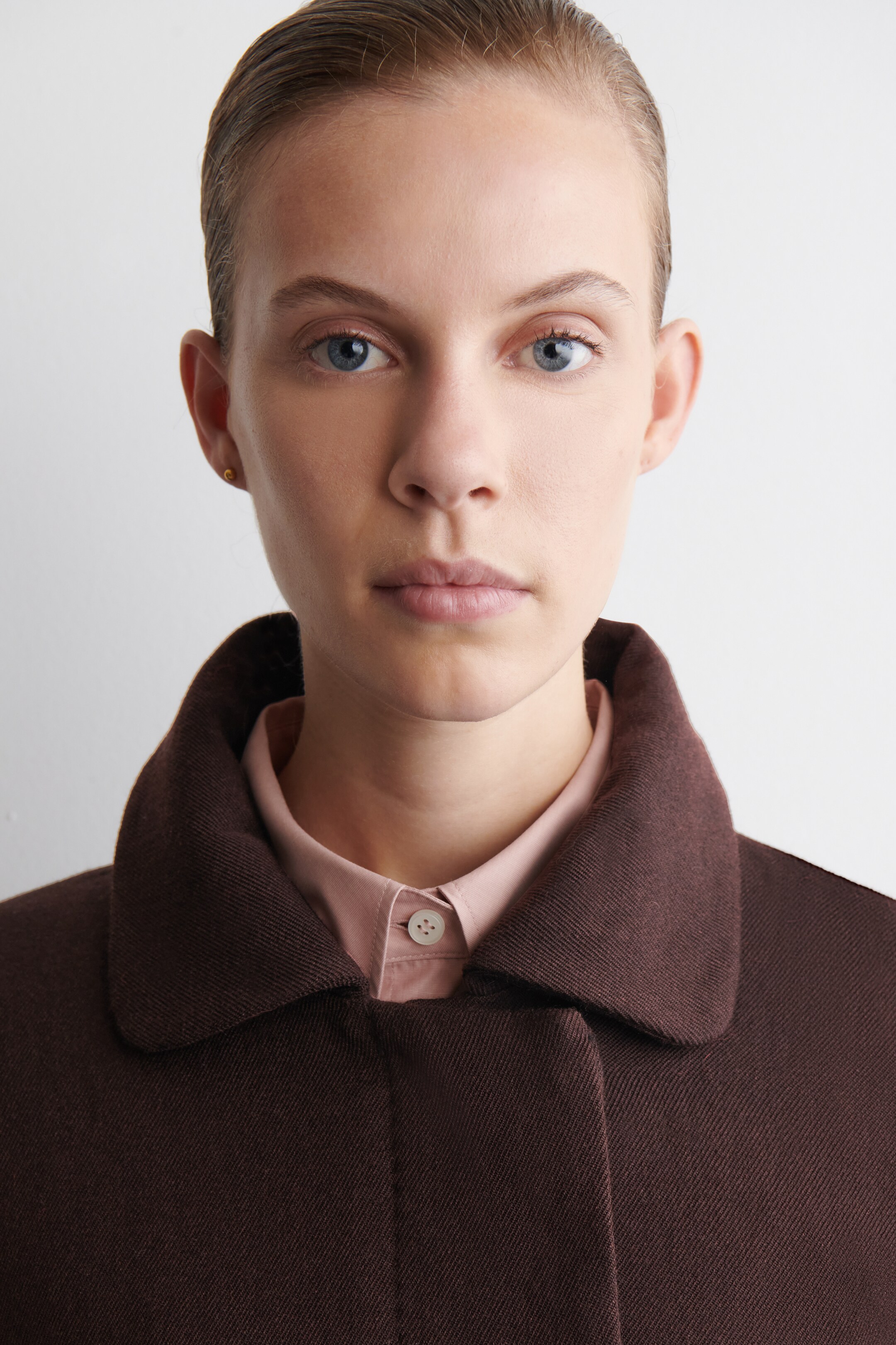 Agrandir l'image: Un manteau brun foncé, avec un col rond de style Peter Pan, est porté. Une chemise rose clair à boutons avec un bouton blanc visible est superposée en dessous.