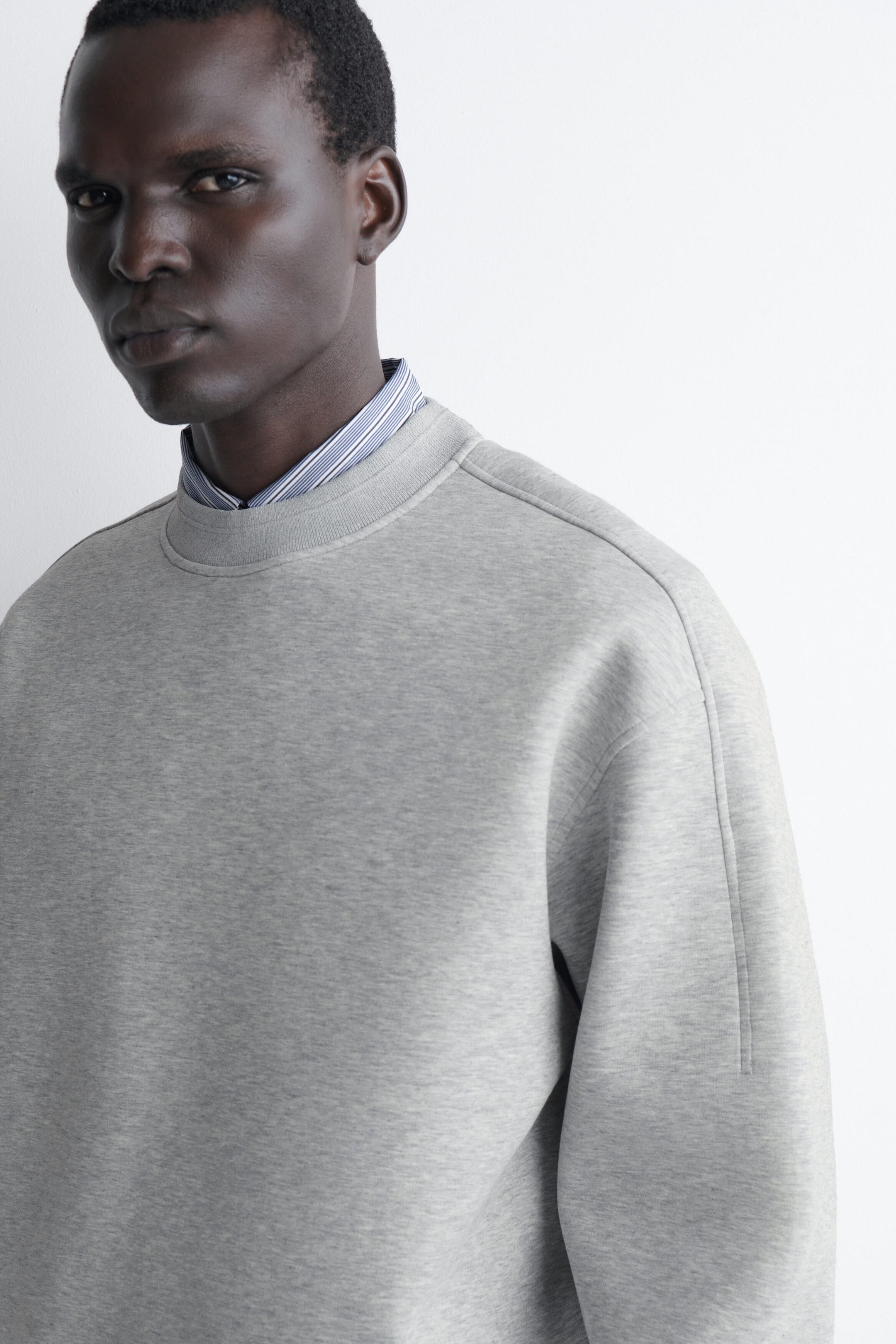 A light grey relaxed scuba sweatshirt is shown in close-up, highlighting its structured fit, ribbed crew neck, and dropped shoulders. The sweatshirt is layered over a blue-and-white striped collared shirt visible at the neckline. A light grey relaxed scuba sweatshirt is shown in close-up, highlighting its structured fit, ribbed crew neck, and dropped shoulders. The sweatshirt is layered over a blue-and-white striped collared shirt visible at the neckline.
