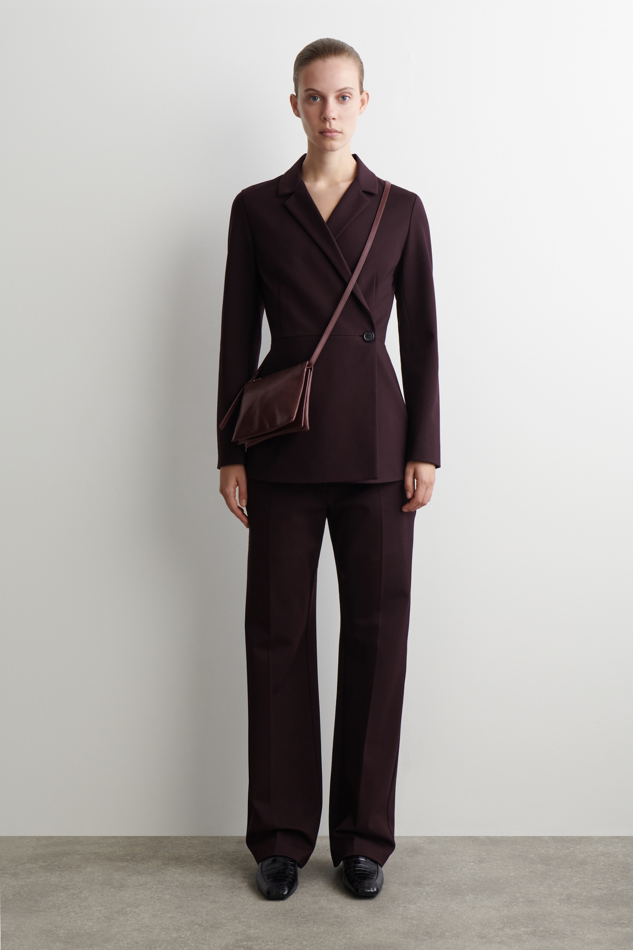 Agrandir l'image: Un tailleur pantalon prune foncé est présenté, avec une veste croisée à revers crantés et une fermeture à un bouton, qui est assortie à un pantalon droit. Un petit sac bandoulière structuré dans une teinte prune foncé similaire est porté par-dessus la veste, et des mocassins noirs complètent l'ensemble.