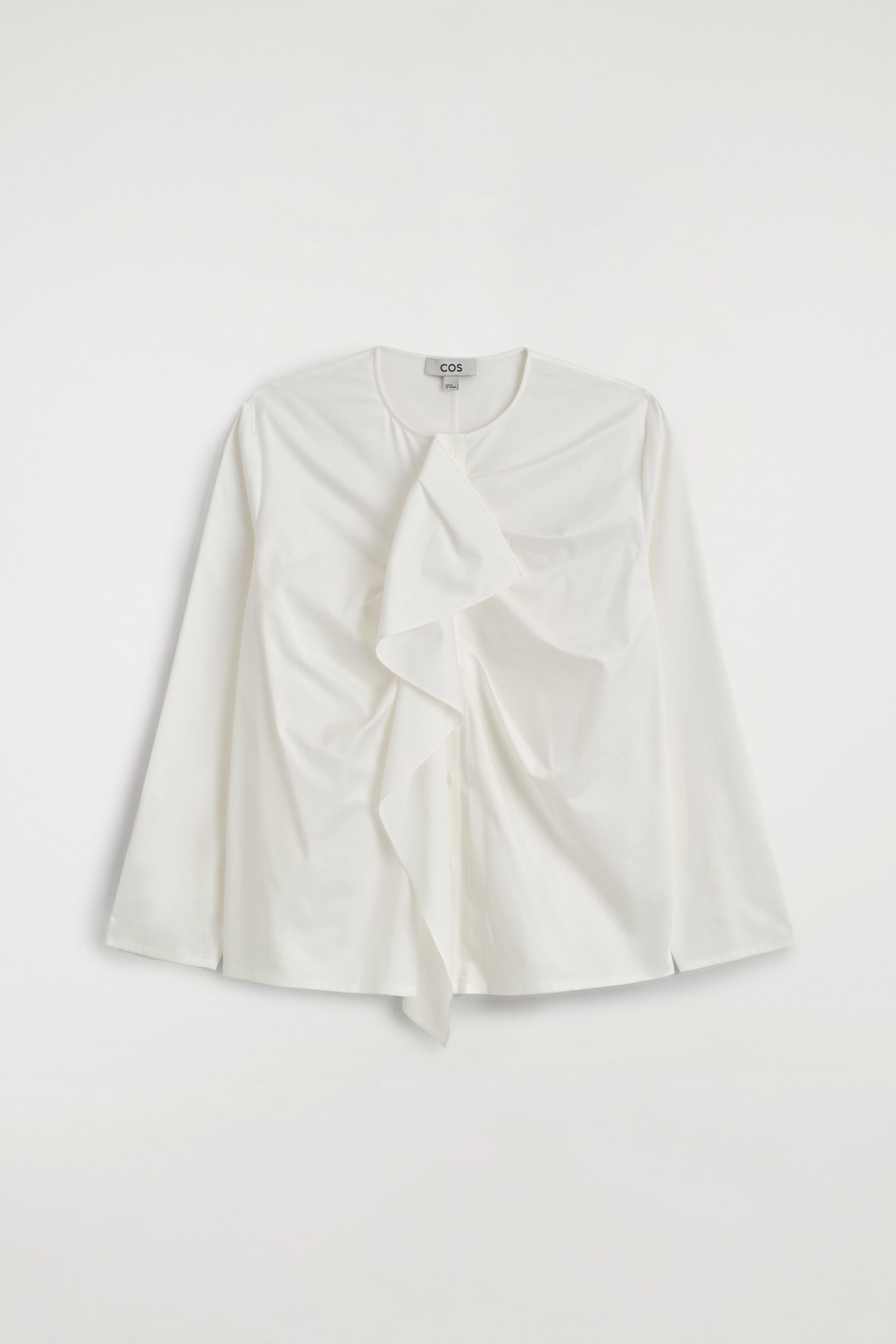 A white, long-sleeved cotton blouse is shown from the front, featuring a collarless round neckline and a soft ruffle cascading down the center placket.