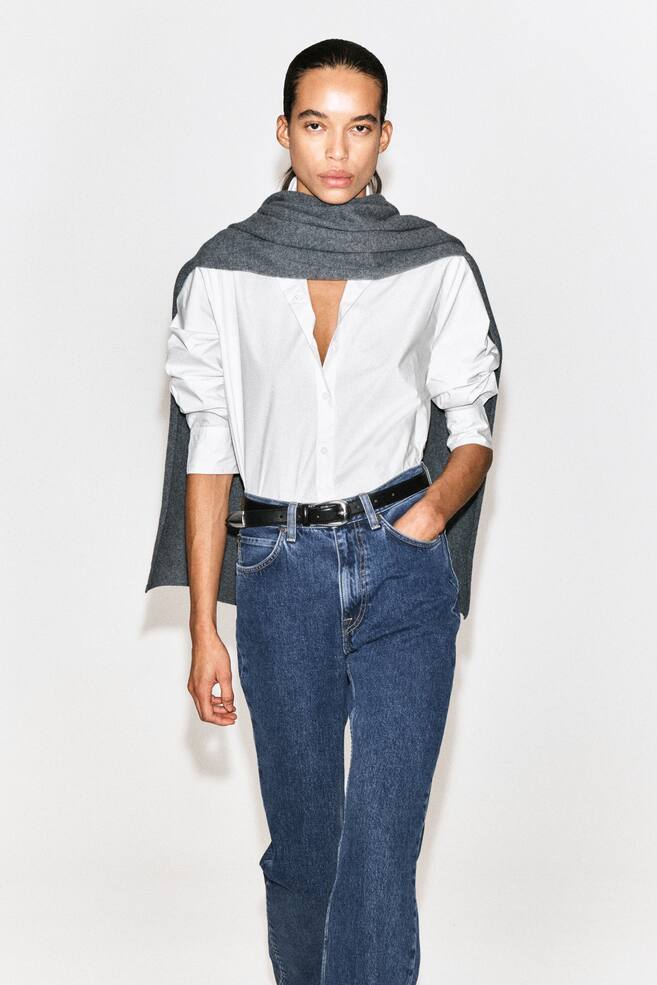 Das klassische Baumwoll-Popeline-Hemd wird von vorne gezeigt und besticht durch ein klares weißes Design mit offenem Kragen und durchgehender Knopfleiste. Es wird in eine blaue Jeans gesteckt getragen, die Ärmel sind hochgekrempelt, während ein grauer Strickpullover locker über die Schultern gelegt ist. - 1
