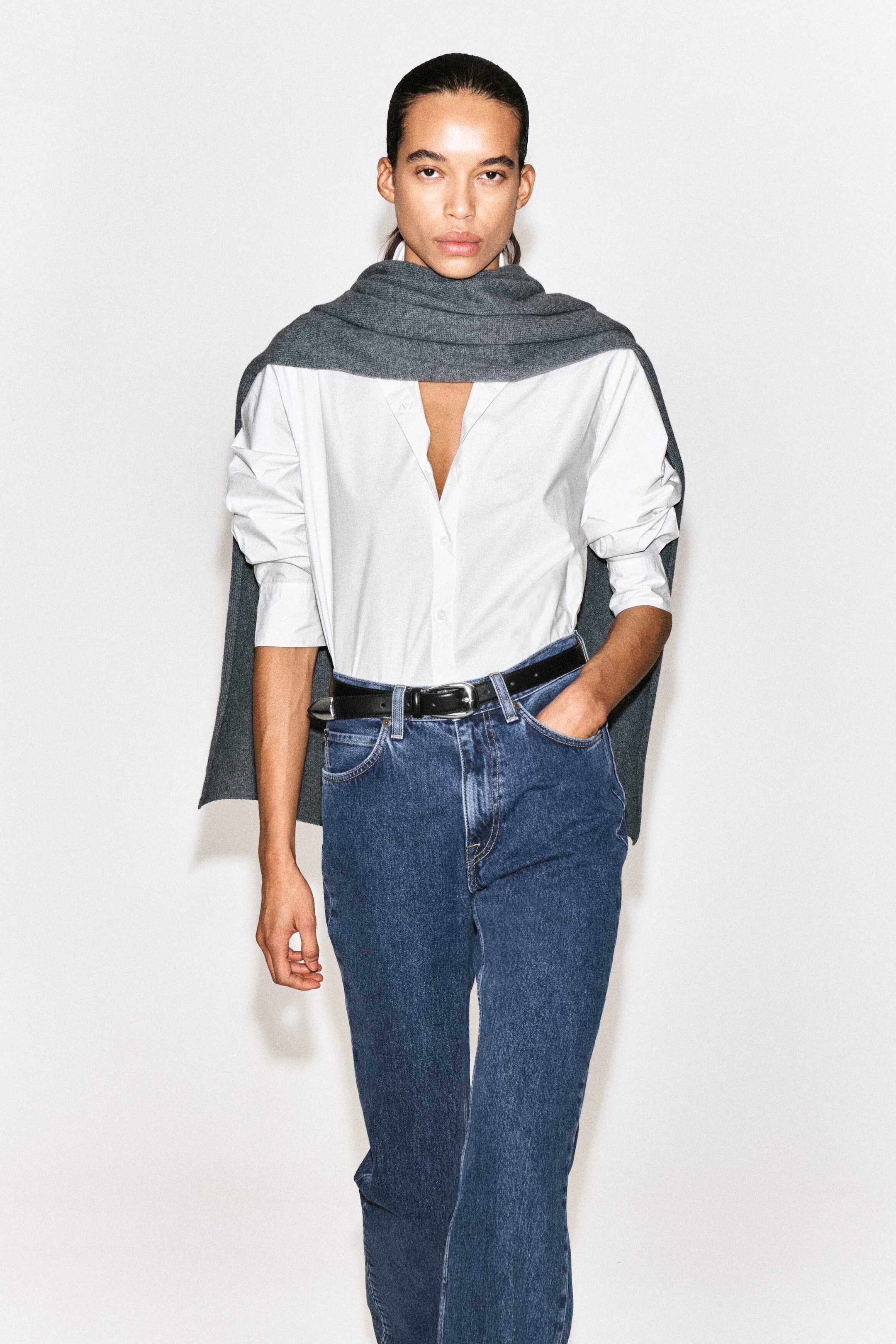 The classic cotton poplin shirt is shown from the front, featuring a crisp white design with an open collar and button-front closure. It is worn tucked into blue jeans with the sleeves rolled up, while a gray knit sweater is draped over the shoulders. The classic cotton poplin shirt is shown from the front, featuring a crisp white design with an open collar and button-front closure. It is worn tucked into blue jeans with the sleeves rolled up, while a gray knit sweater is draped over the shoulders.