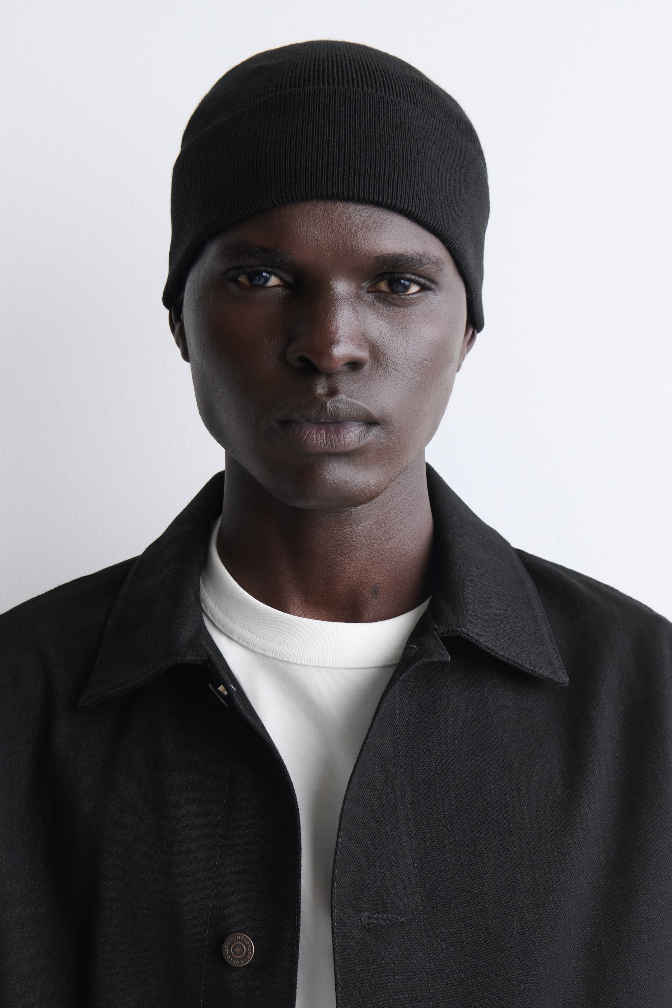 Zobacz większy obraz: Czarna czapka typu beanie o drobnym splocie, wykonana w bezszwowej konstrukcji i z podwiniętym brzegiem, noszona przez modela zwróconego przodem do aparatu.