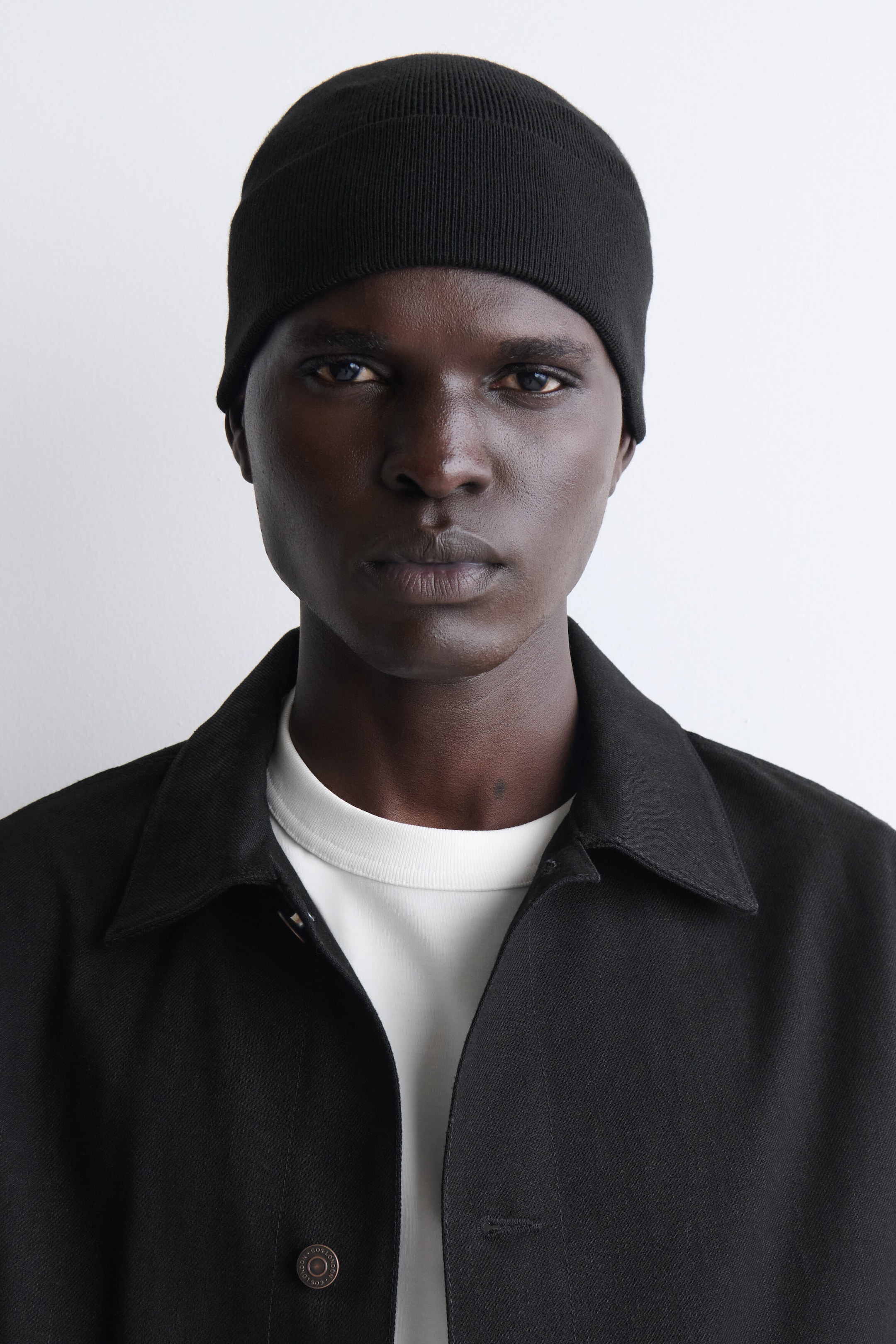 A black fine-knit beanie with seamless construction and a fold-up brim is being worn by a model facing forward.