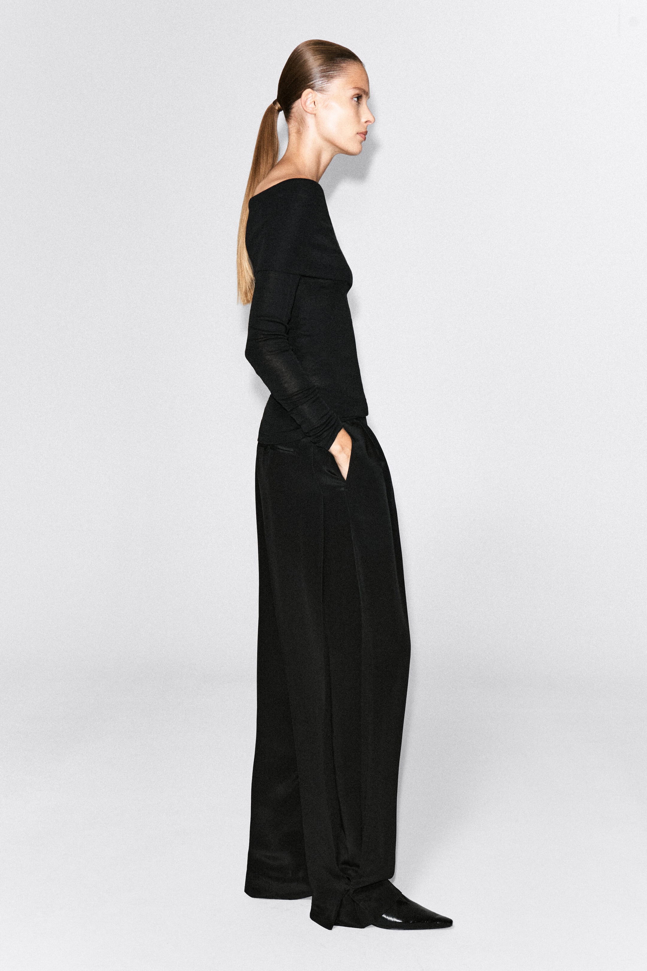 The model is wearing an off-the-shoulder black merino wool top featuring a slim fit, long sleeves, and a folded neckline. The top is styled with wide-leg black trousers.