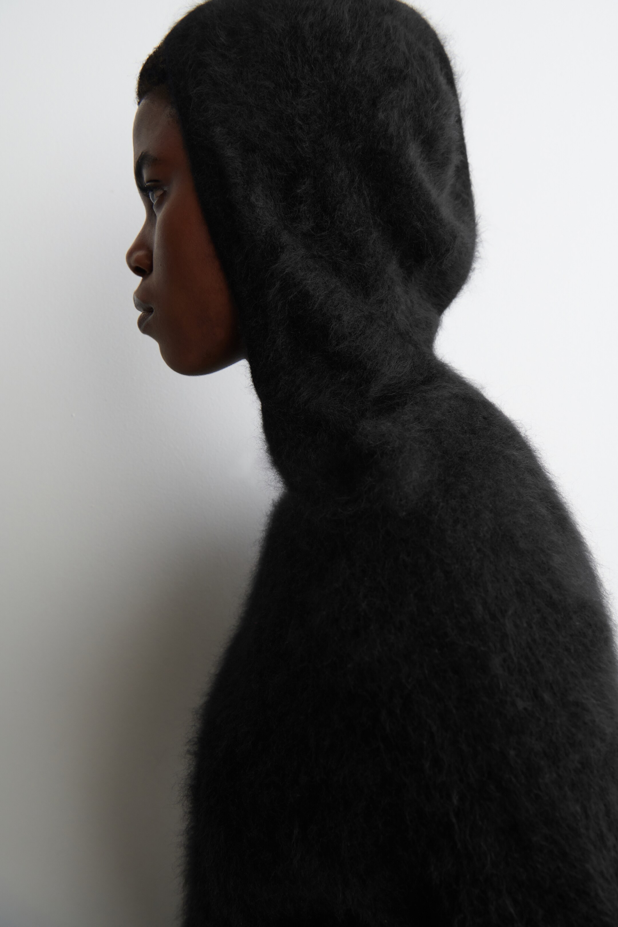 View larger image: A black, hooded garment with a visibly fuzzy, long-pile texture is covering the head and neck, shown from a side profile. The garment is fitting snugly around the head and extending downwards over the neck and shoulders.