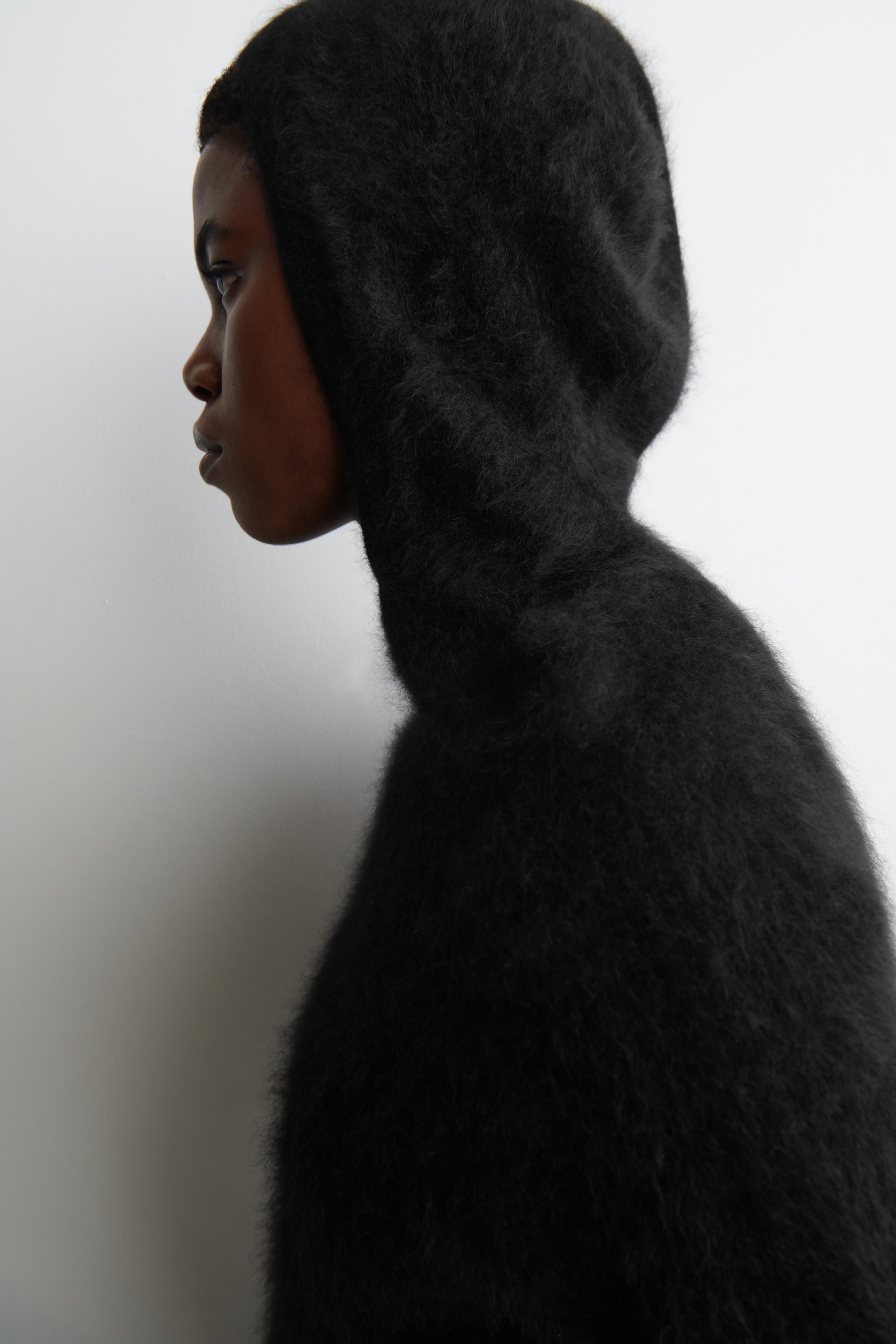 View larger image: A black, hooded garment with a visibly fuzzy, long-pile texture is covering the head and neck, shown from a side profile. The garment is fitting snugly around the head and extending downwards over the neck and shoulders.