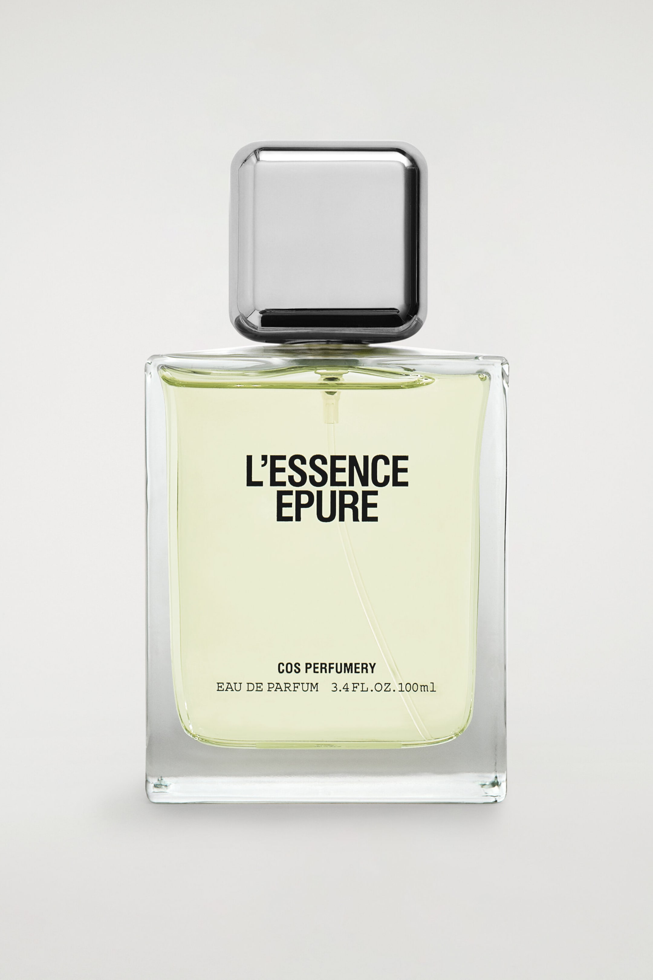 A rectangular glass bottle of COS Perfumery "L'Essence Epure" Eau de Parfum stands upright in a front-facing view, containing a pale yellow liquid. The bottle features a metallic, square-shaped silver cap with rounded corners and displays black printed text on the front reading "L'ESSENCE EPURE", "COS PERFUMERY", and "EAU DE PARFUM 3.4 FL.OZ. 100ml".