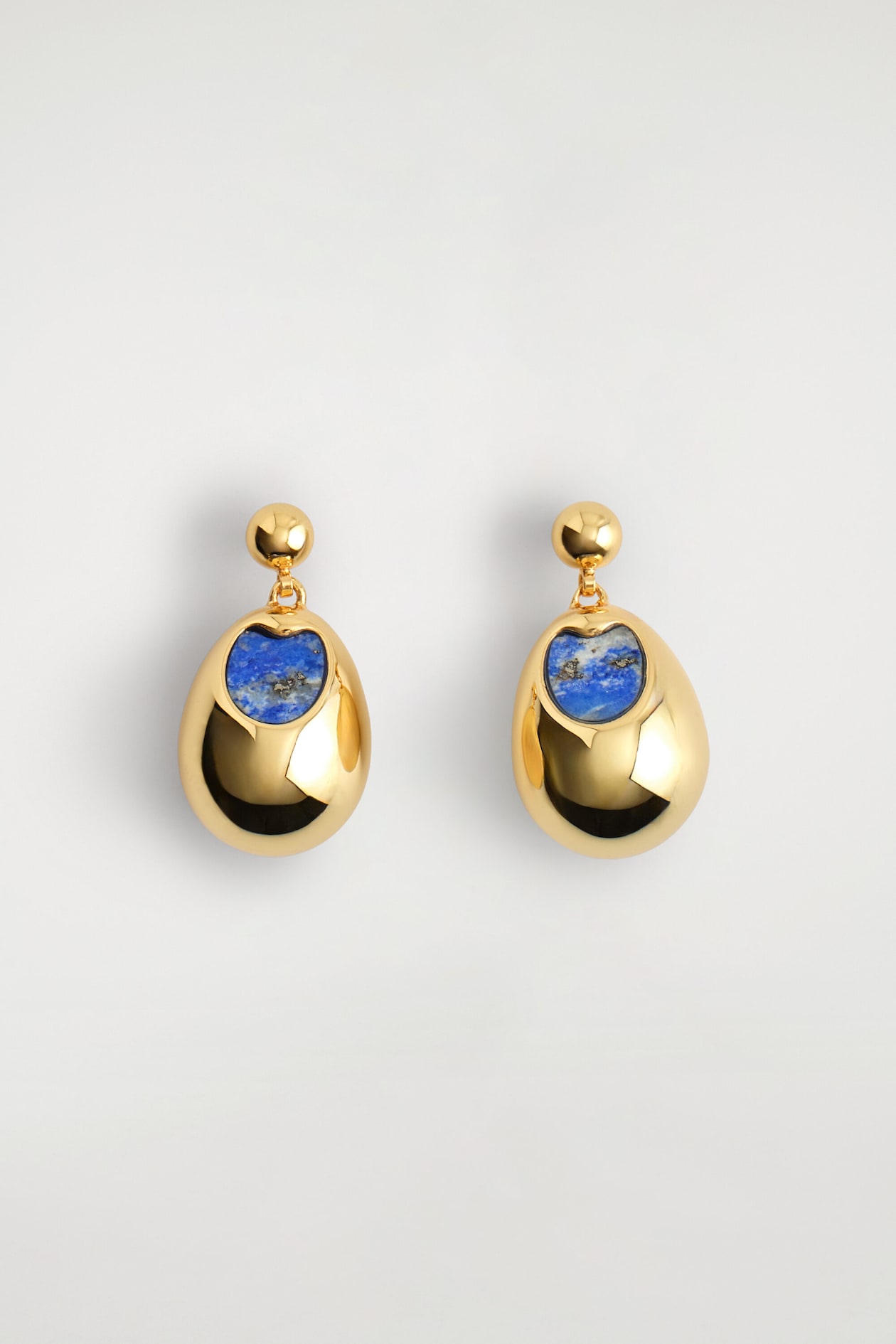 Une paire de boucles d’oreilles à pendants avec détail en pierre est présentée de face, avec une finition ton doré hautement polie. Chaque boucle d’oreille présente un petit clou sphérique relié à un pendentif en forme de goutte bombée, incrusté d’une pierre bleue texturée.