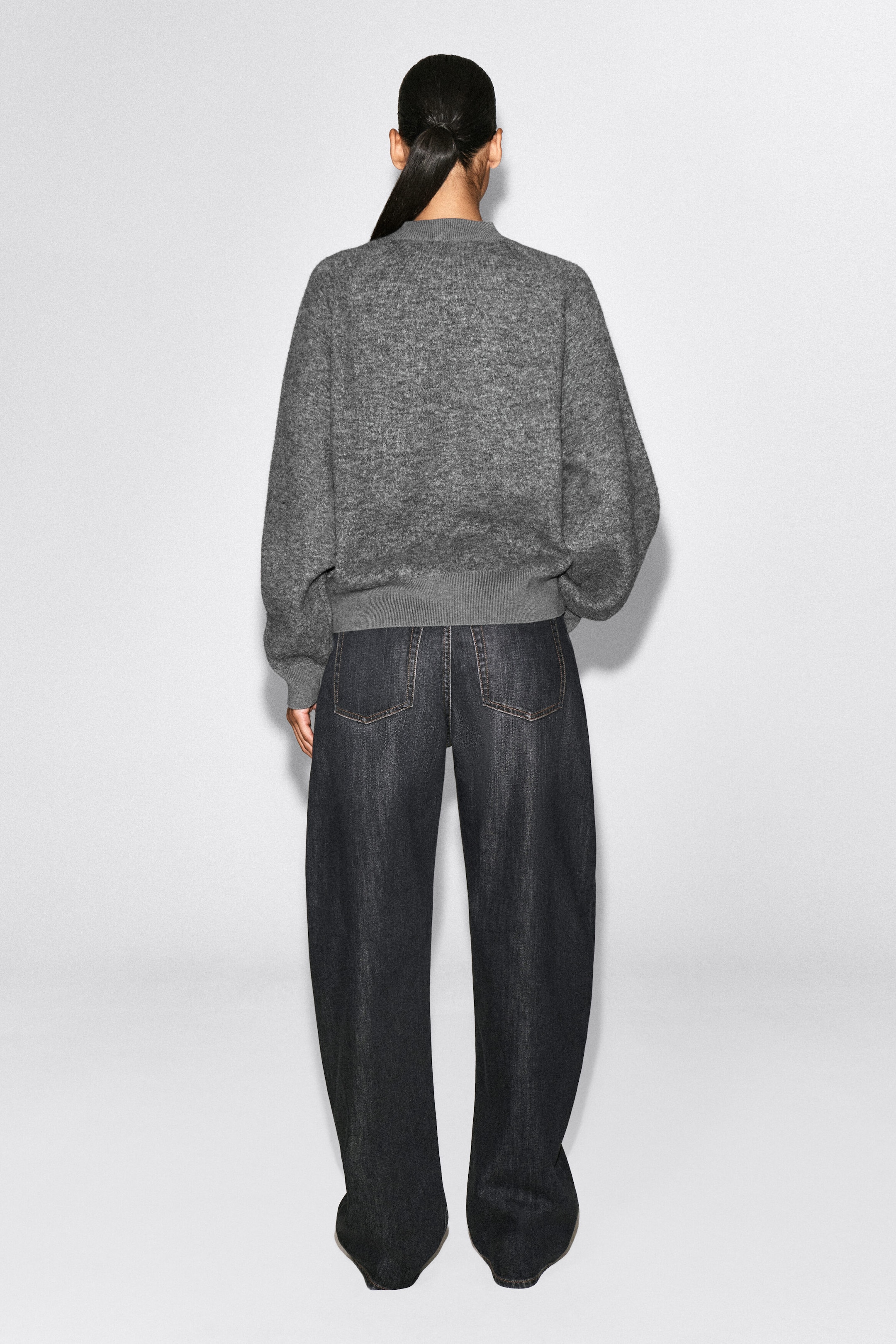 A textured gray knit sweater is shown from the back, featuring a relaxed fit, dropped shoulders, and ribbed hem and cuffs. The sweater is paired with dark gray wide-leg jeans featuring classic rear patch pockets. A textured gray knit sweater is shown from the back, featuring a relaxed fit, dropped shoulders, and ribbed hem and cuffs. The sweater is paired with dark gray wide-leg jeans featuring classic rear patch pockets.