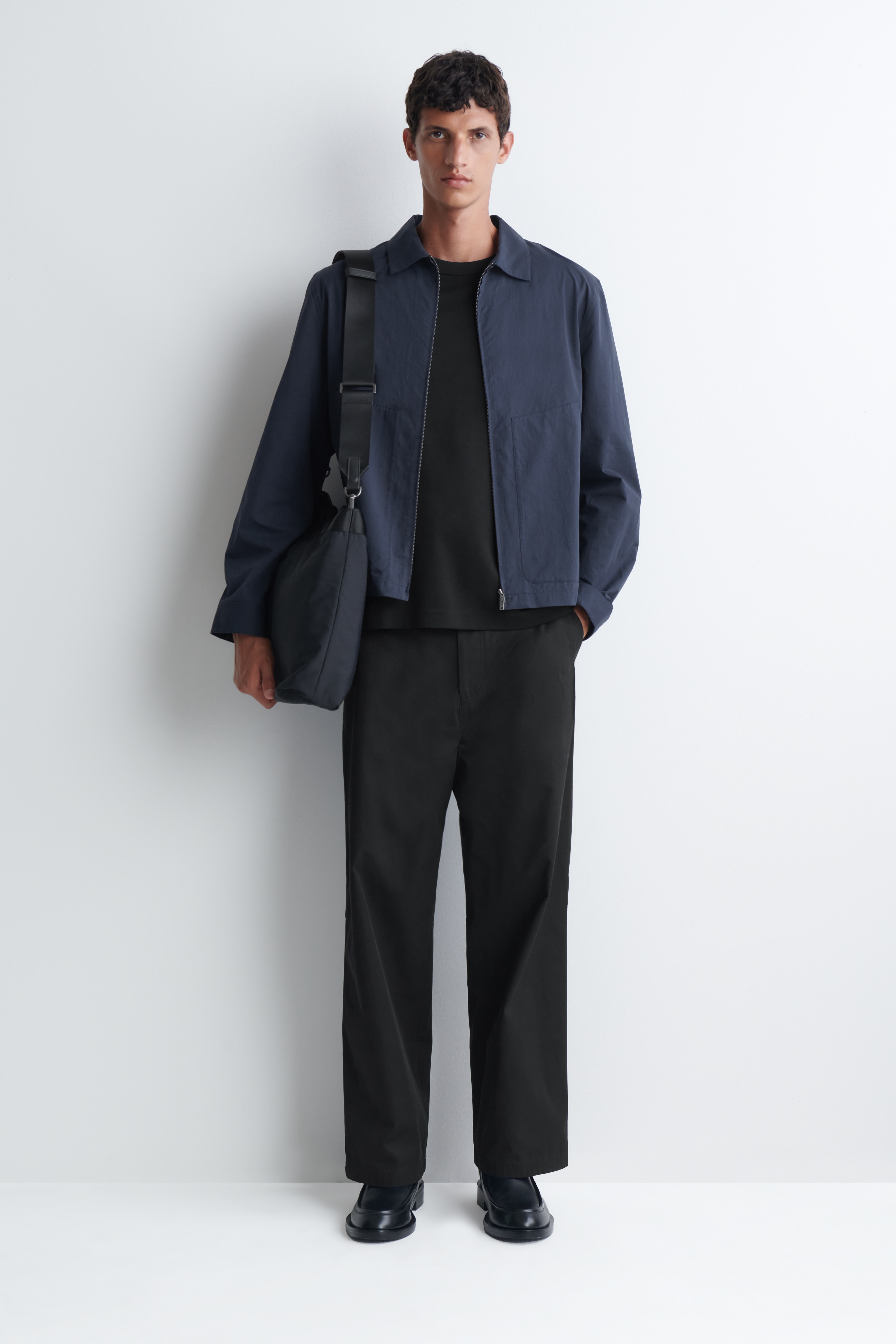 A black fabric bag is shown from the front, featuring a slouchy rectangular shape with dual top handles and a long adjustable shoulder strap attached with metal clasps.
