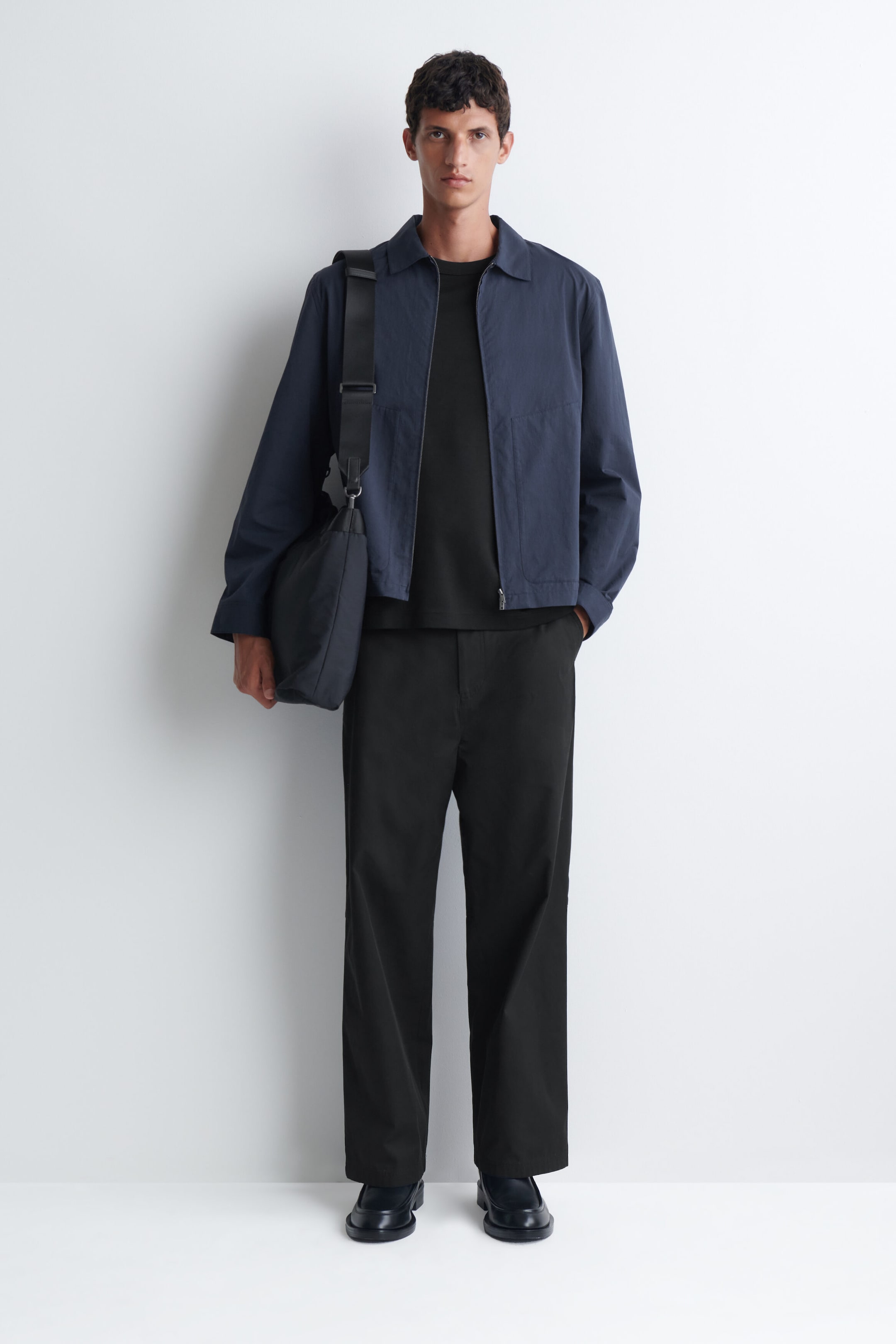 A navy blue zip-up shirt-jacket is being worn by the model over a black t-shirt and wide-leg trousers. The jacket features a point collar and a silver-tone front zipper, and is styled with a matching dark shoulder bag carried over one shoulder.