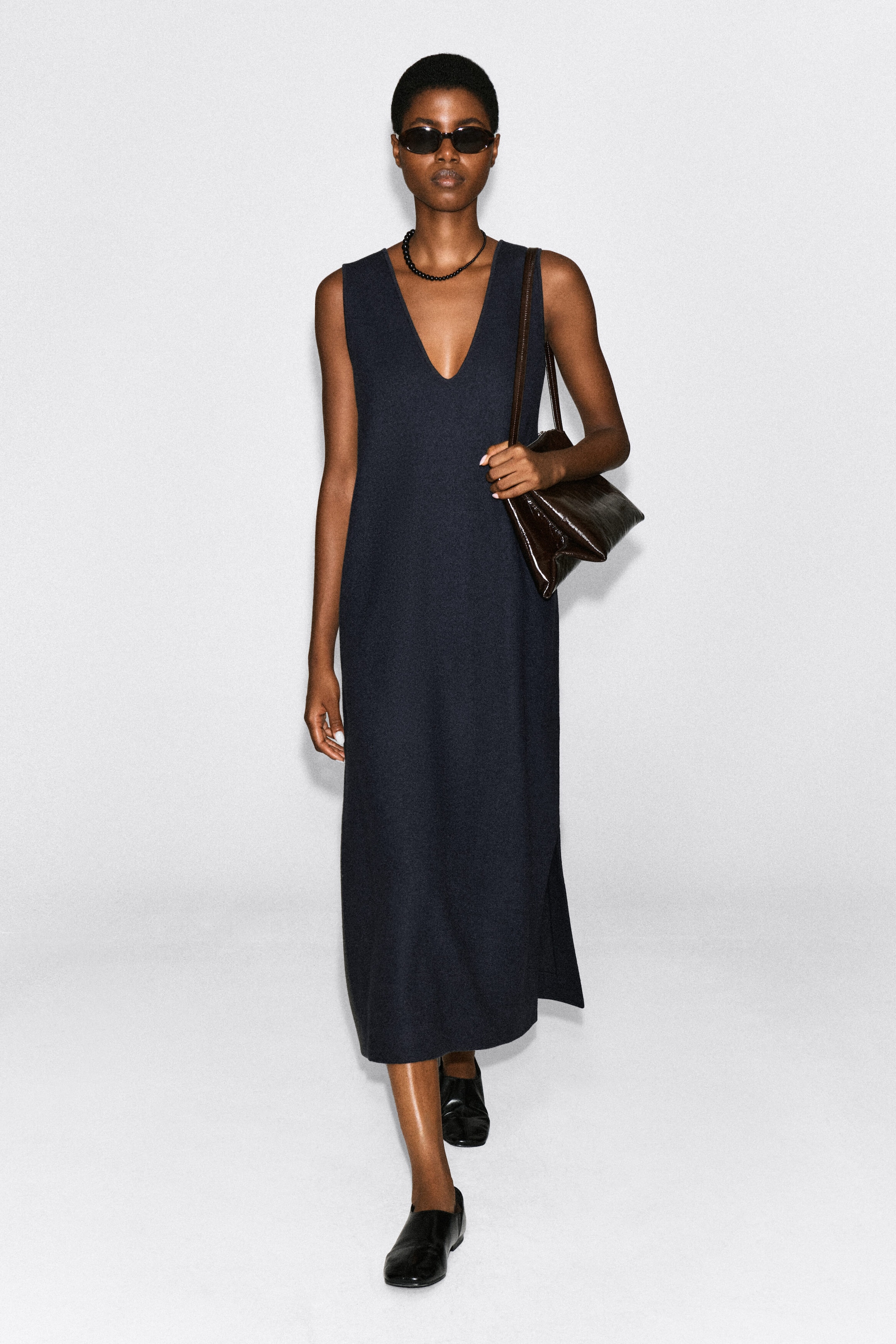 A dark navy boiled wool-blend midi dress with a deep V-neckline and sleeveless cut is worn by the model. The dress is styled with a dark brown textured leather shoulder bag, black sunglasses, a beaded necklace, and black leather slip-on shoes. A dark navy boiled wool-blend midi dress with a deep V-neckline and sleeveless cut is worn by the model. The dress is styled with a dark brown textured leather shoulder bag, black sunglasses, a beaded necklace, and black leather slip-on shoes.