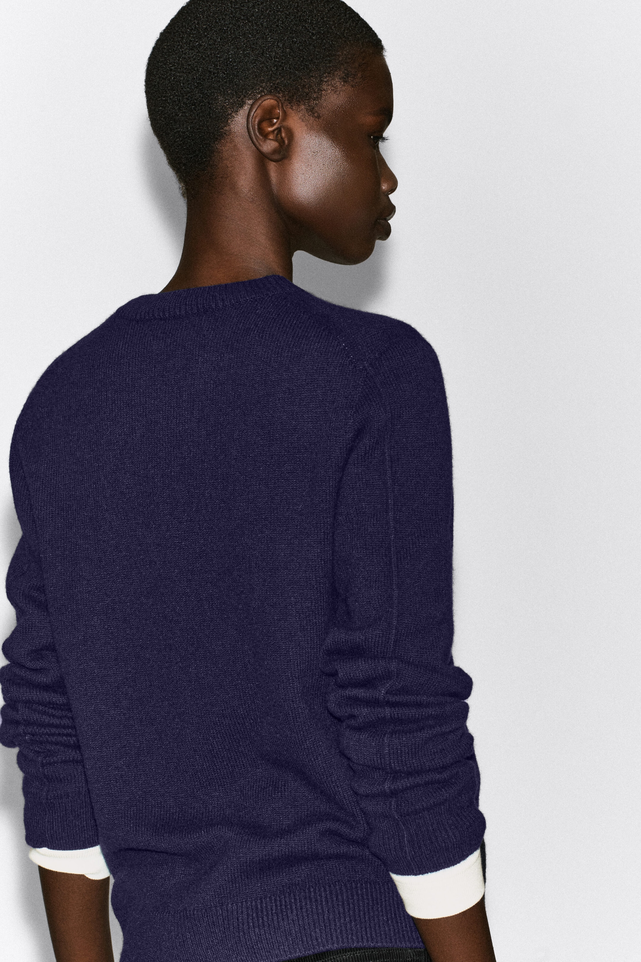 Un pull en cachemire bleu marine foncé est porté par le mannequin, vu de dos pour mettre en valeur son col rond côtelé et son ourlet côtelé. Des poignets blancs d’un sous-vêtement superposé dépassent sous les manches longues du pull. Un pull en cachemire bleu marine foncé est porté par le mannequin, vu de dos pour mettre en valeur son col rond côtelé et son ourlet côtelé. Des poignets blancs d’un sous-vêtement superposé dépassent sous les manches longues du pull.