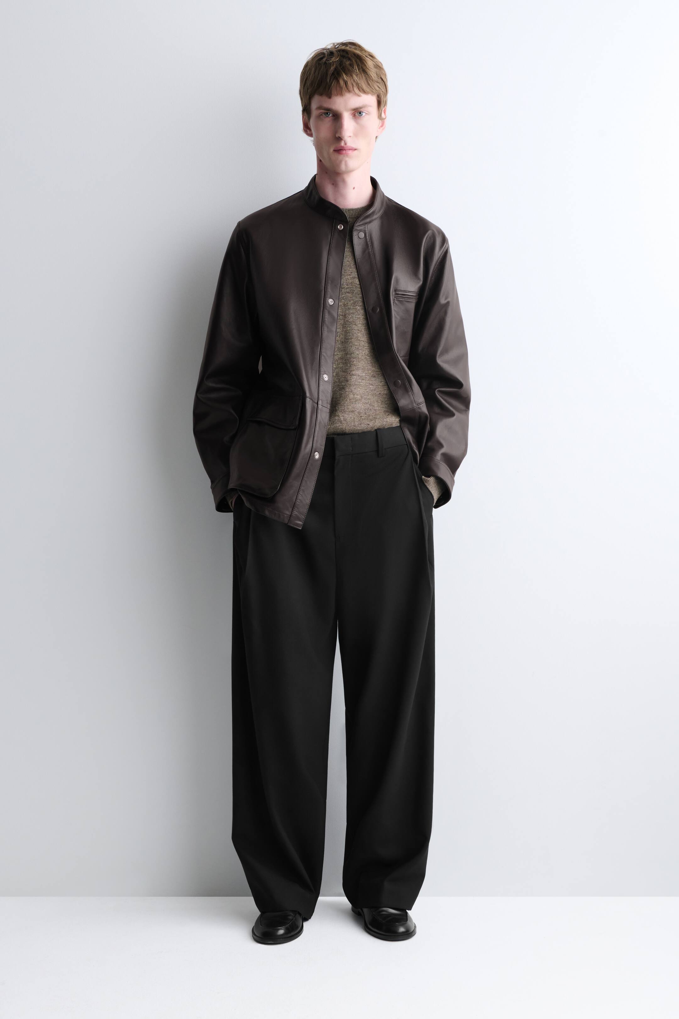 View larger image: A brown leather band-collar jacket, featuring a front button fastening, two lower patch pockets, and an upper chest pocket, is worn over a beige textured jumper, which is paired with black pleated wide-leg trousers and black plain-toe loafers.