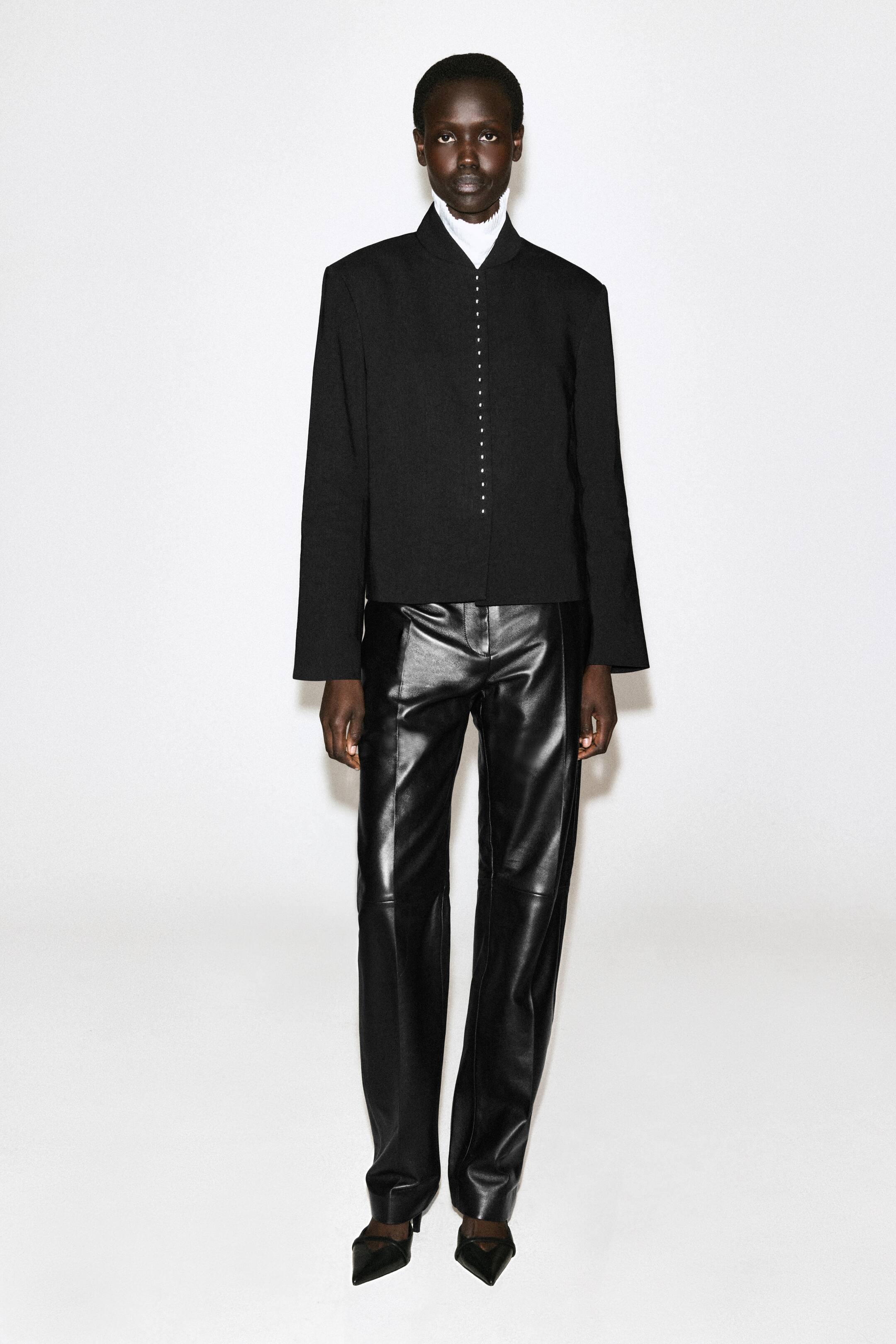 View larger image: A black Mandarin-collar jacket with a column of small buttons down the front is being layered over a white high-neck top, paired with black straight-leg leather trousers featuring prominent front seams, and black pointed-toe shoes.
