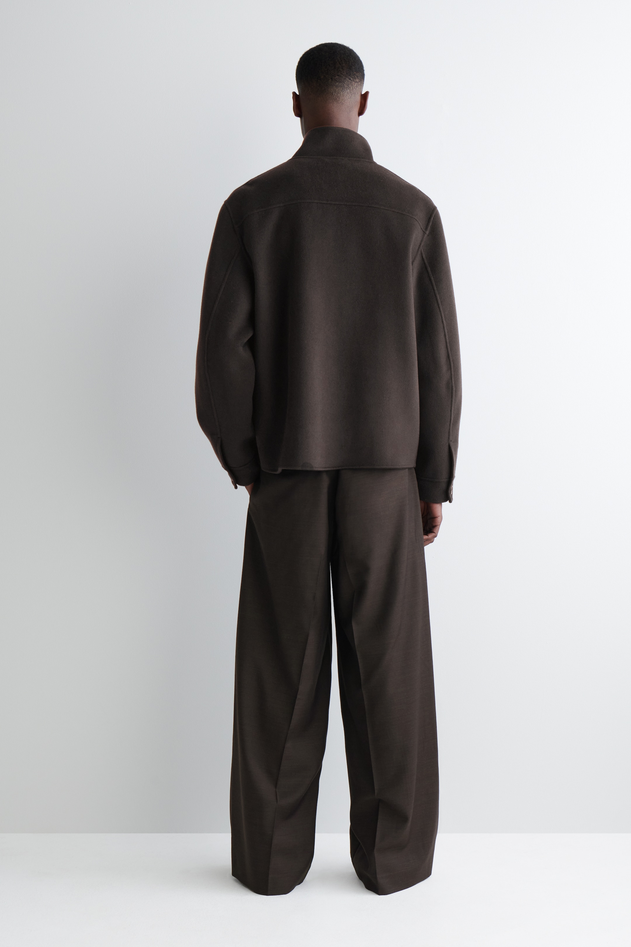 Grösseres Bild ansehen: Eine dunkelbraune Harrington-Jacke wird von dem Modell getragen, das steht. Die Jacke hat einen Stehkragen und geknöpfte Manschetten und wird mit passenden dunkelbraunen Hosen mit weitem Bein gestylt.
