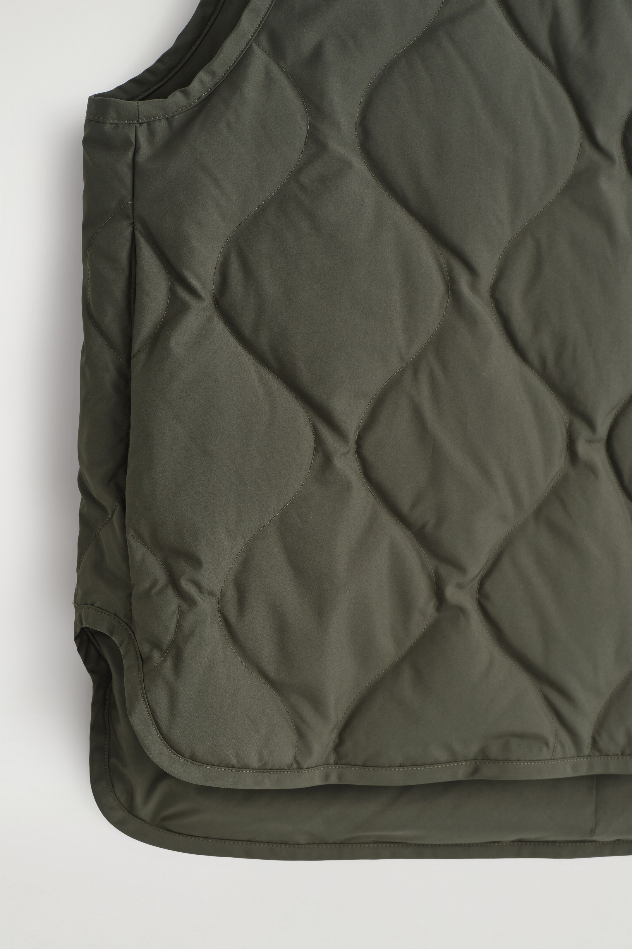 Agrandir l'image: Un gilet matelassé vert olive est exposé de face, avec un motif matelassé ondulé et un ourlet incurvé.