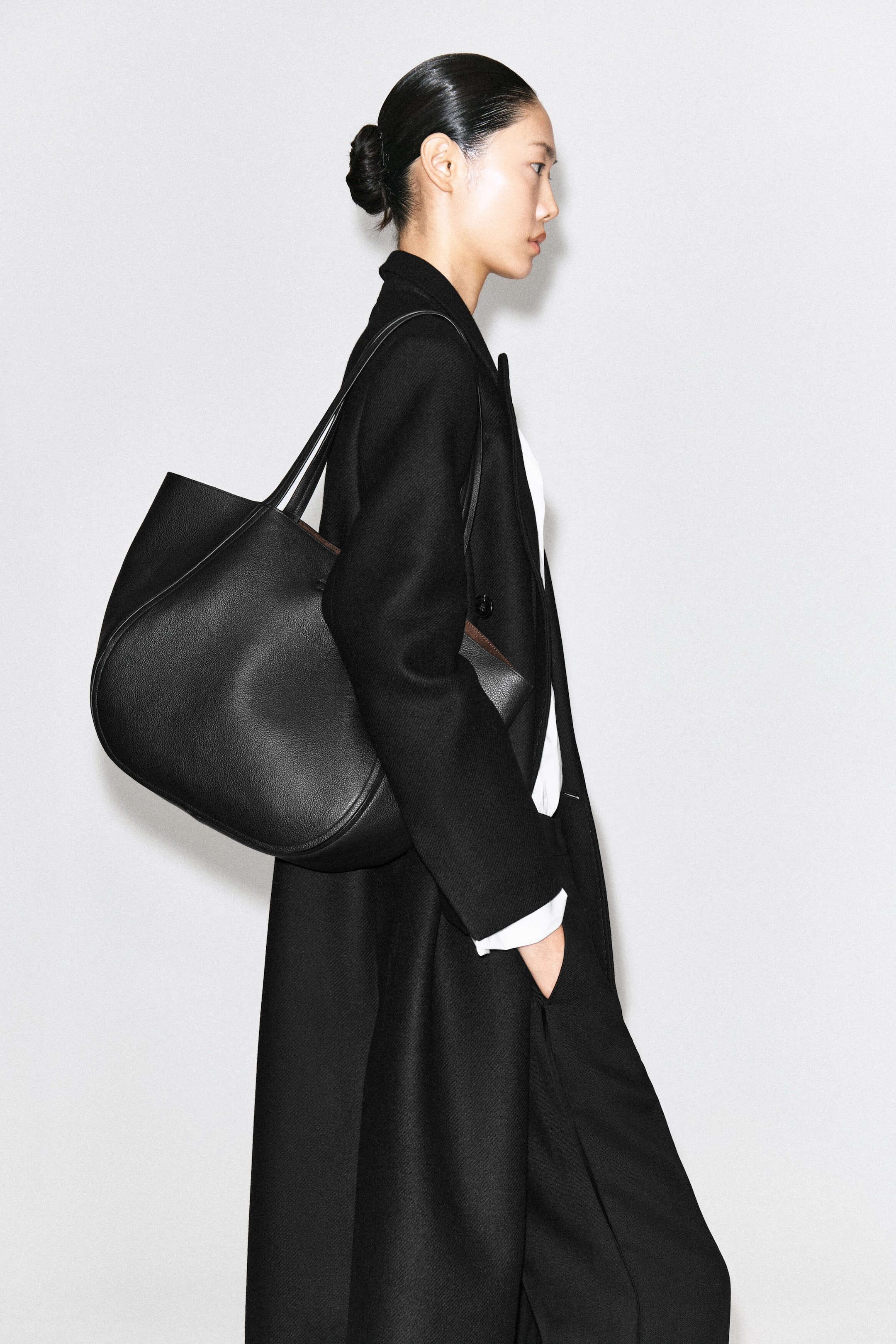 A black grained leather shopper bag with a curved silhouette and dual shoulder straps is being carried over the model’s shoulder. The bag rests against a long black overcoat worn by the model. A black grained leather shopper bag with a curved silhouette and dual shoulder straps is being carried over the model’s shoulder. The bag rests against a long black overcoat worn by the model.