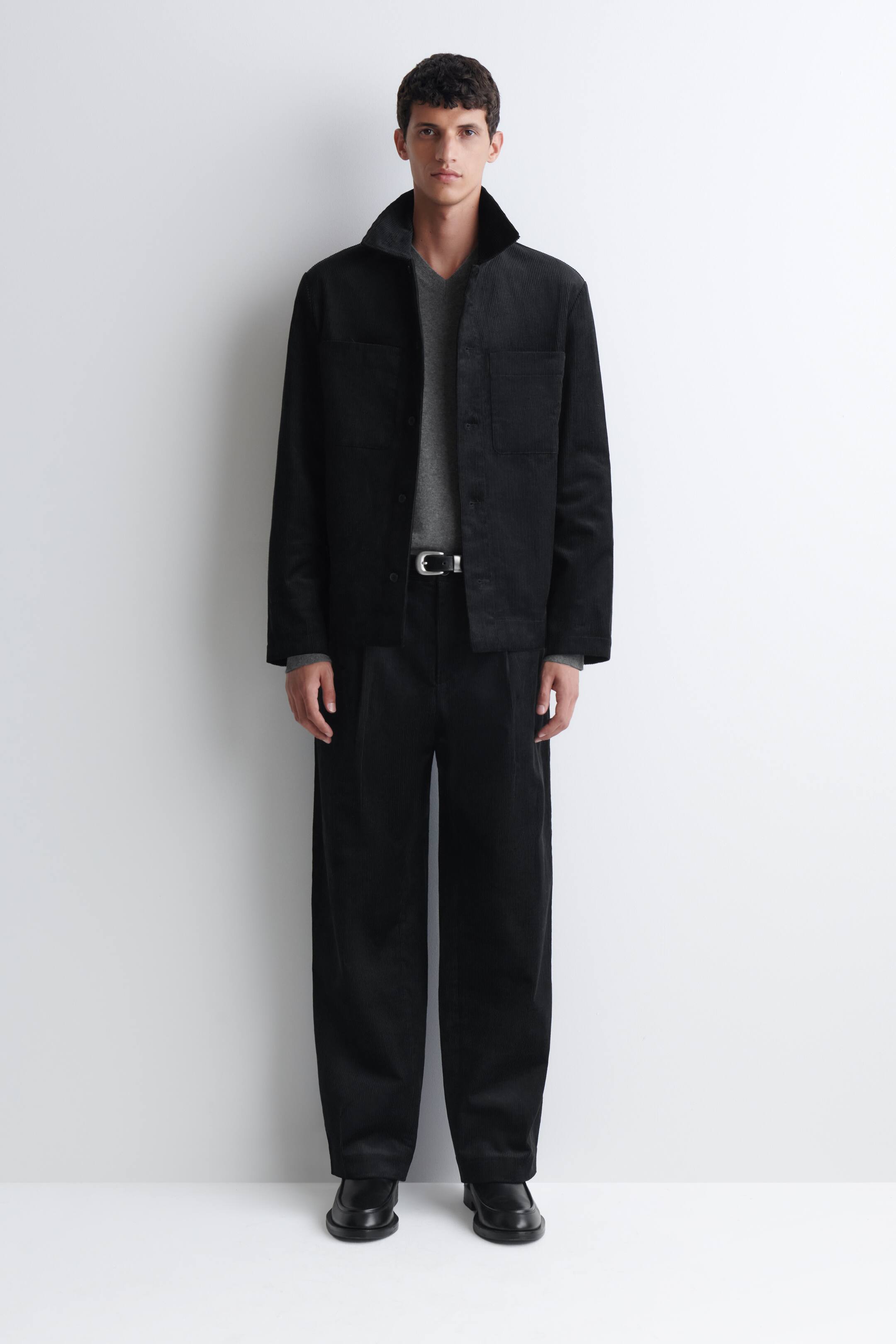 View larger image: A black cotton corduroy overshirt is worn unbuttoned by the model, featuring a pointed collar, front button fastening, and patch chest pockets. The overshirt is layered over a grey V-neck top and paired with matching black trousers.