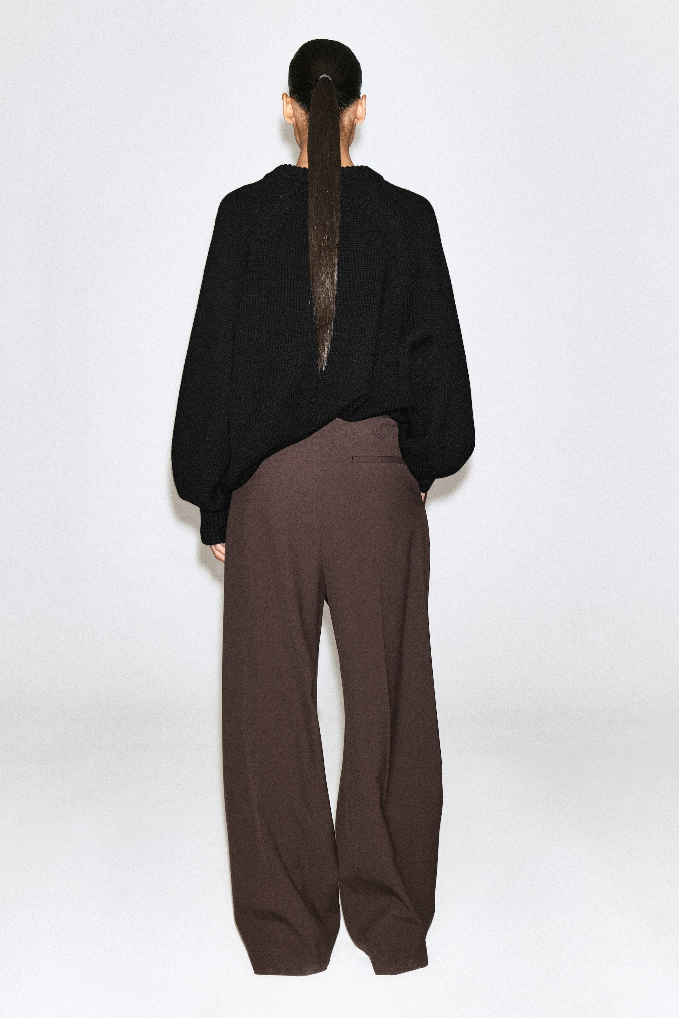 This black chunky-knit sweater features a relaxed silhouette with ribbed cuffs, shown from the back as it drapes over the waistband of dark brown, wide-leg trousers. The trousers feature a loose, flowing fit and a single back welt pocket.